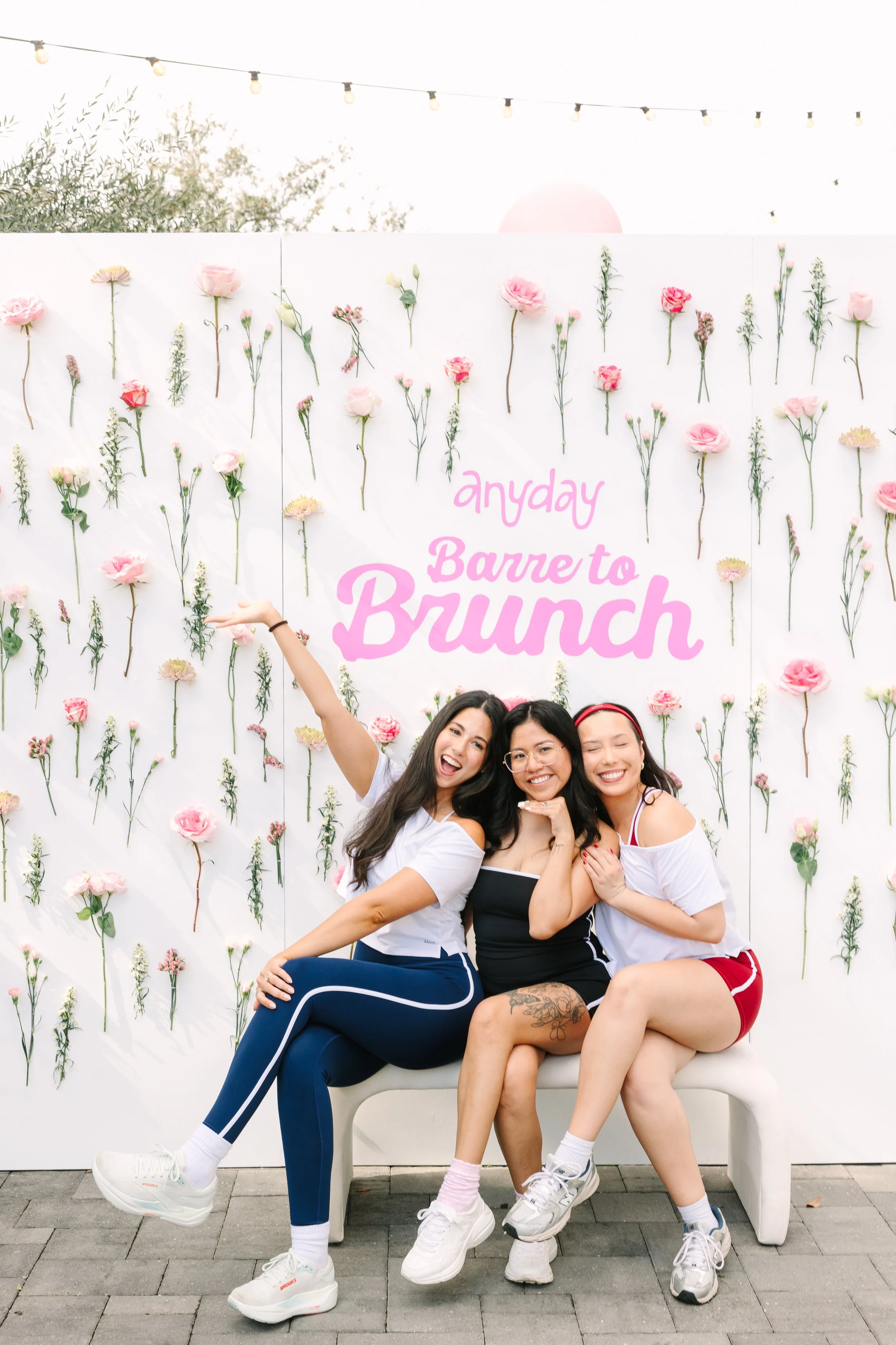 Three women sitting on a white bench smiling and posing in front of a floral backdrop with the text 'anyday Barre to Brunch' at an outdoor event. Barre to Brunch with Jenna Palek