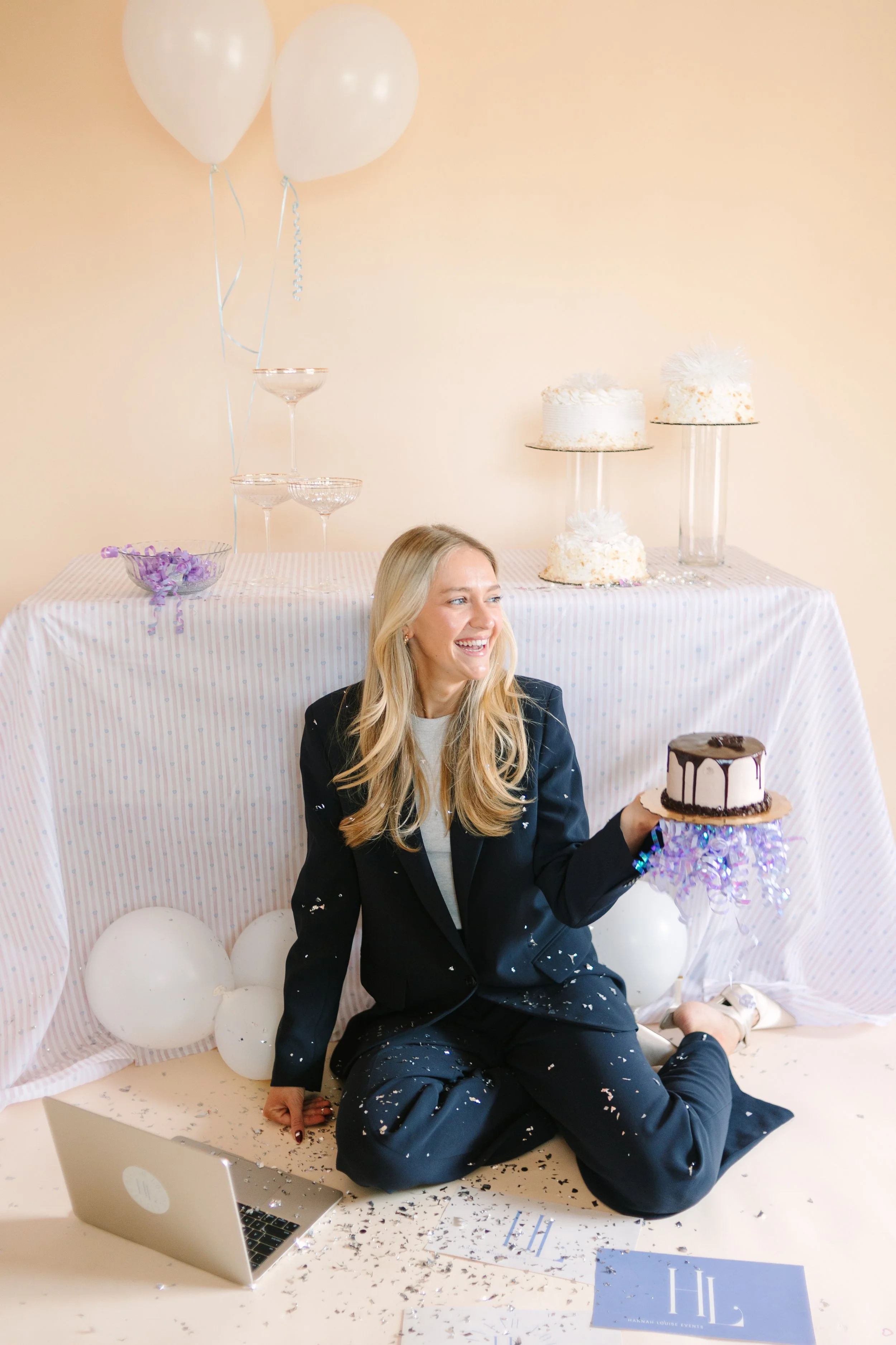 A woman in a black suit sitting on the floor, holding a small cake, surrounded by balloons and confetti. In the background, there are decorated cakes on a table and a laptop on the floor. Event Planner