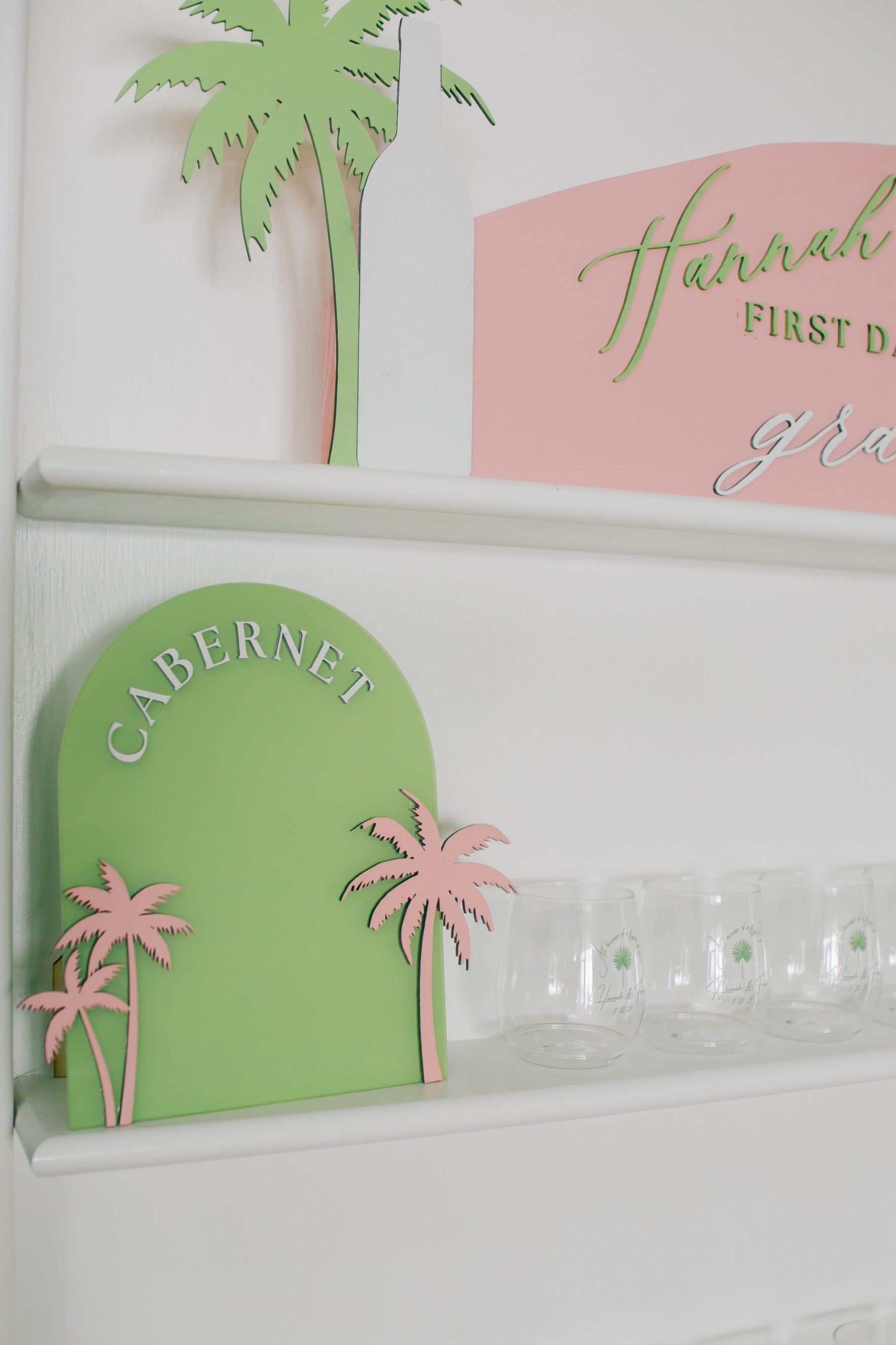 Decorative shelves with signs reading "CABERNET" and "Hannah First Day" with pink and green tropical palm tree cutouts and a row of clear glasses.