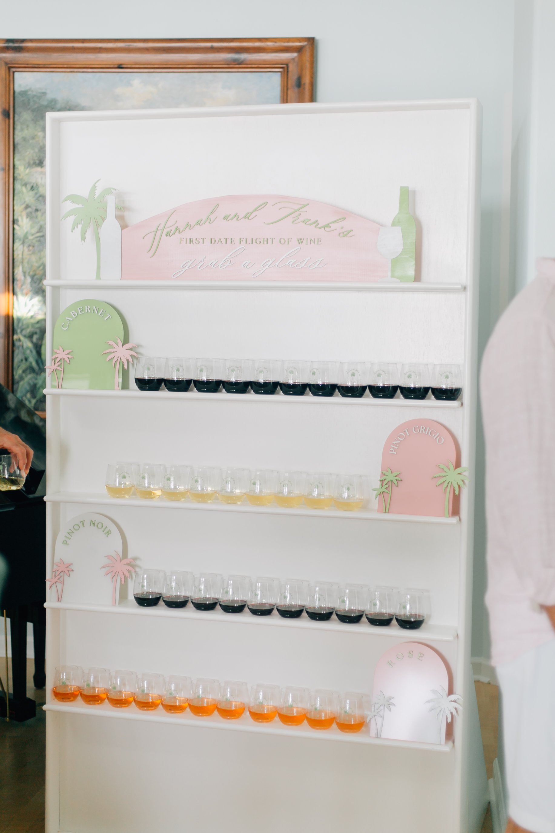 A wine tasting display with multiple levels of glasses filled with different types of wine, labeled with signs such as 'Cabernet', 'Pinot Grigio', 'Pinot Noir', and 'Rose', with a decorative backdrop featuring palm trees and the text 'Hannah and Fran