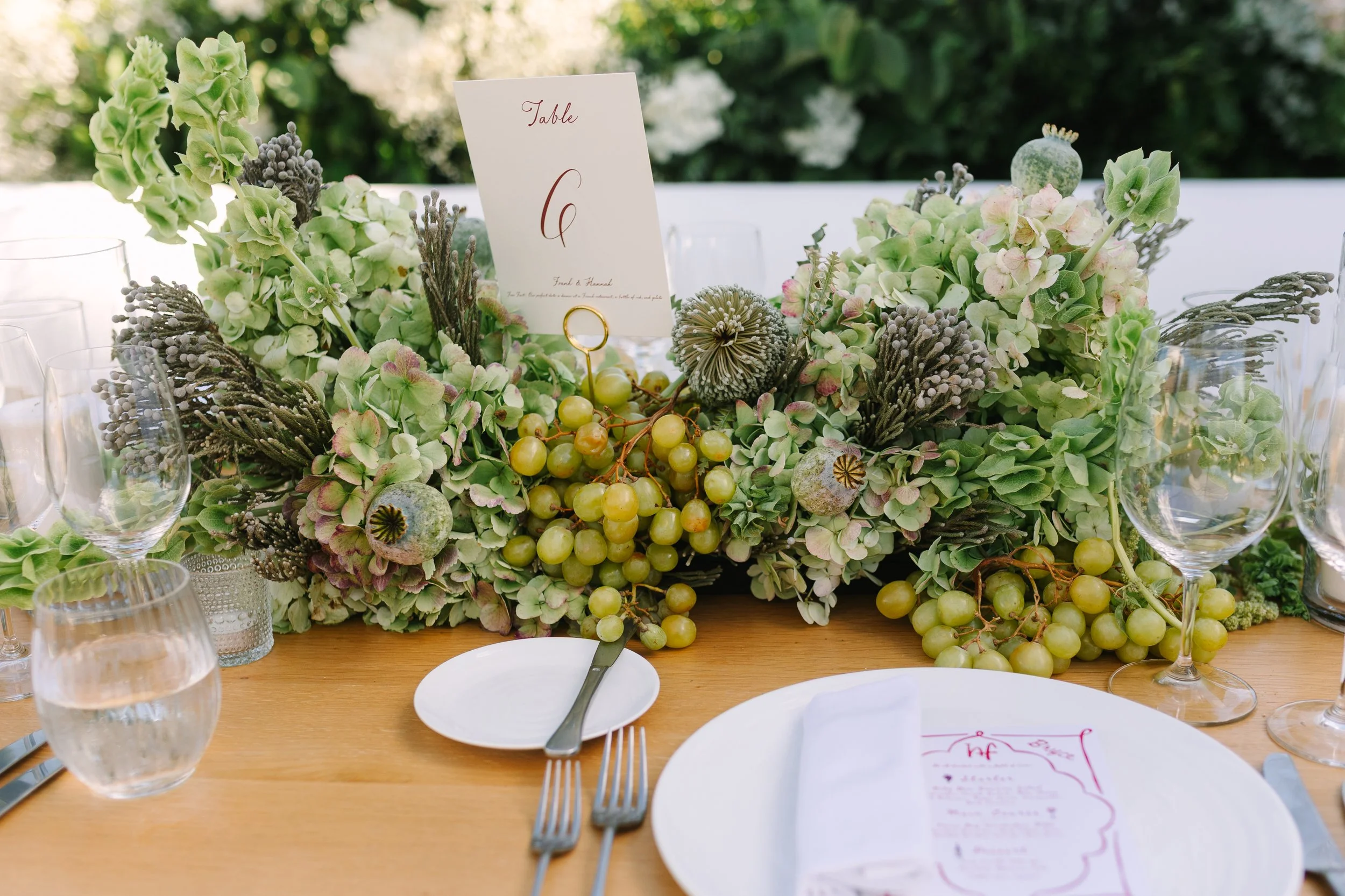 A beautifully decorated wedding table with a floral centerpiece of green and purple flowers, green grapes, and decorative elements, along with glassware, a place setting, and a table number card.