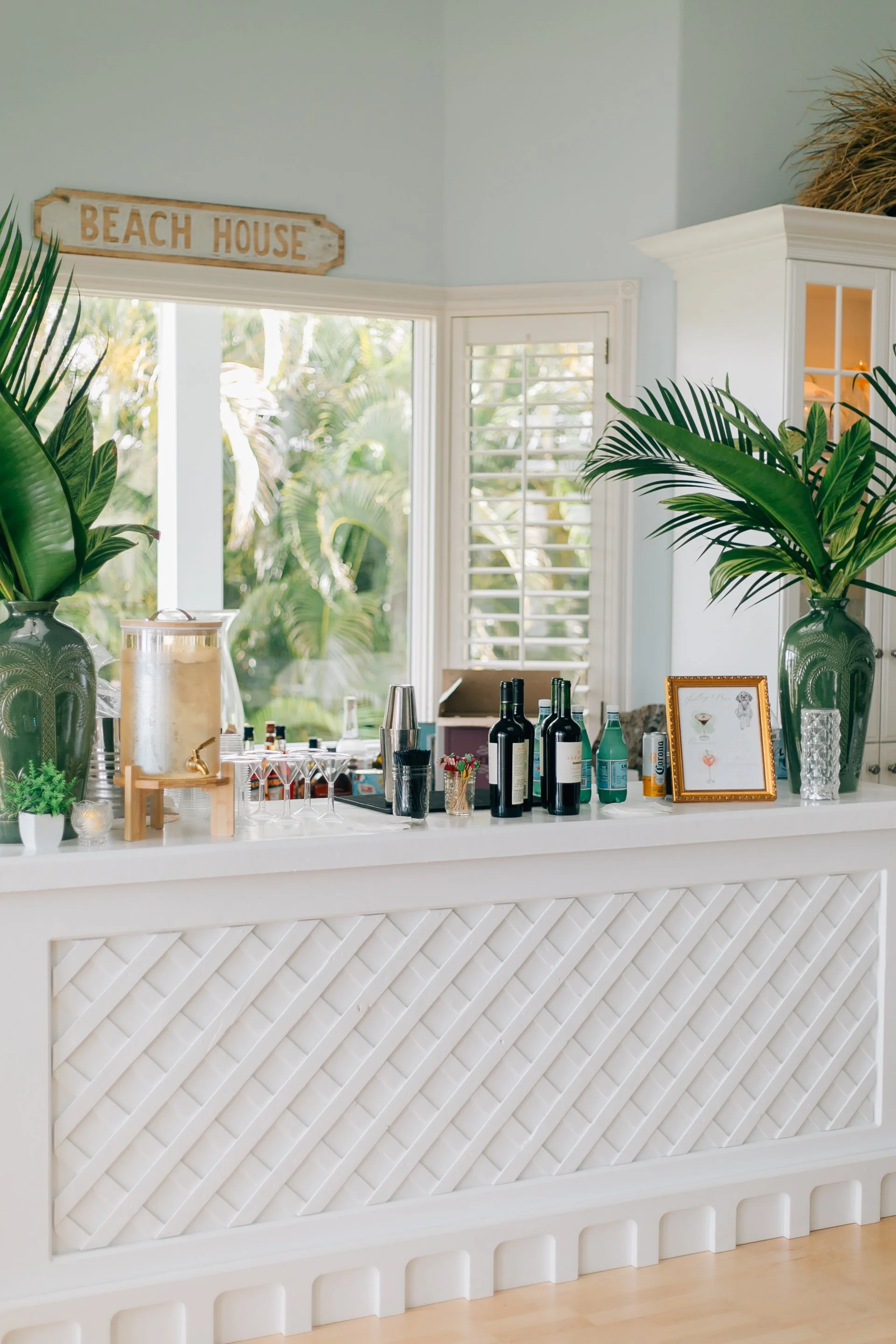 A white bar counter with tropical plants in vases, wine bottles, framed photo, and drinkware, set in a bright room with large windows and a 'Beach House' sign. Engagement Party Sarasota, FL