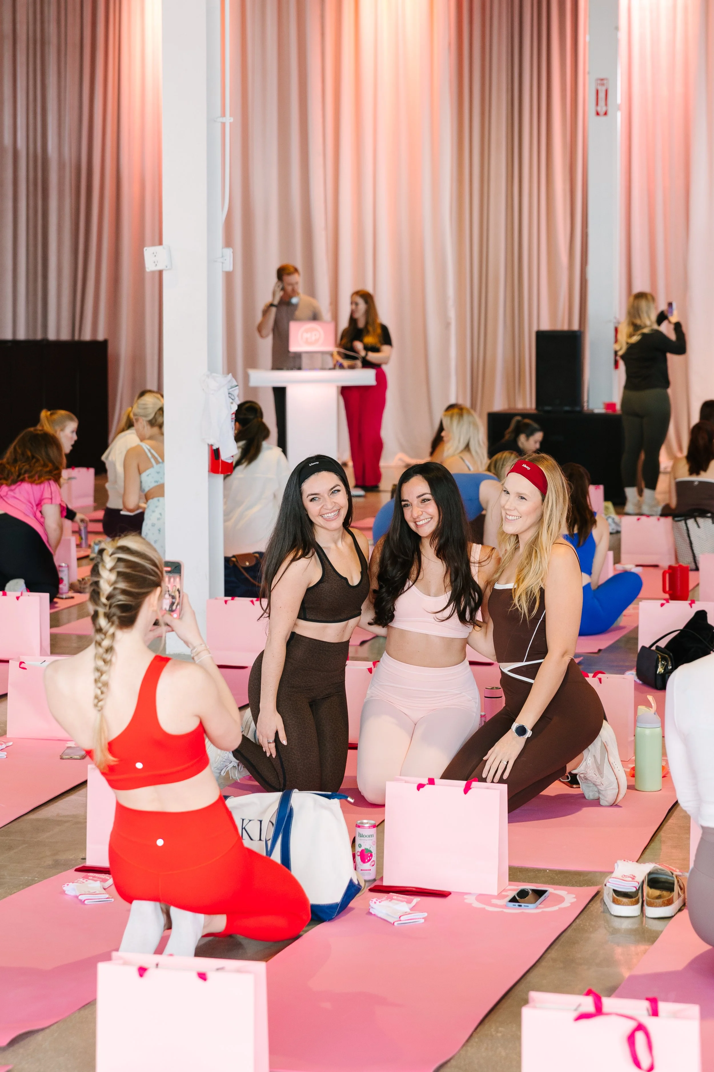 Women participating in a fitness event, some taking pictures, with three women posing in the center in front of a pink backdrop and fitness mats. Barre to Brunch with Jenna Palek