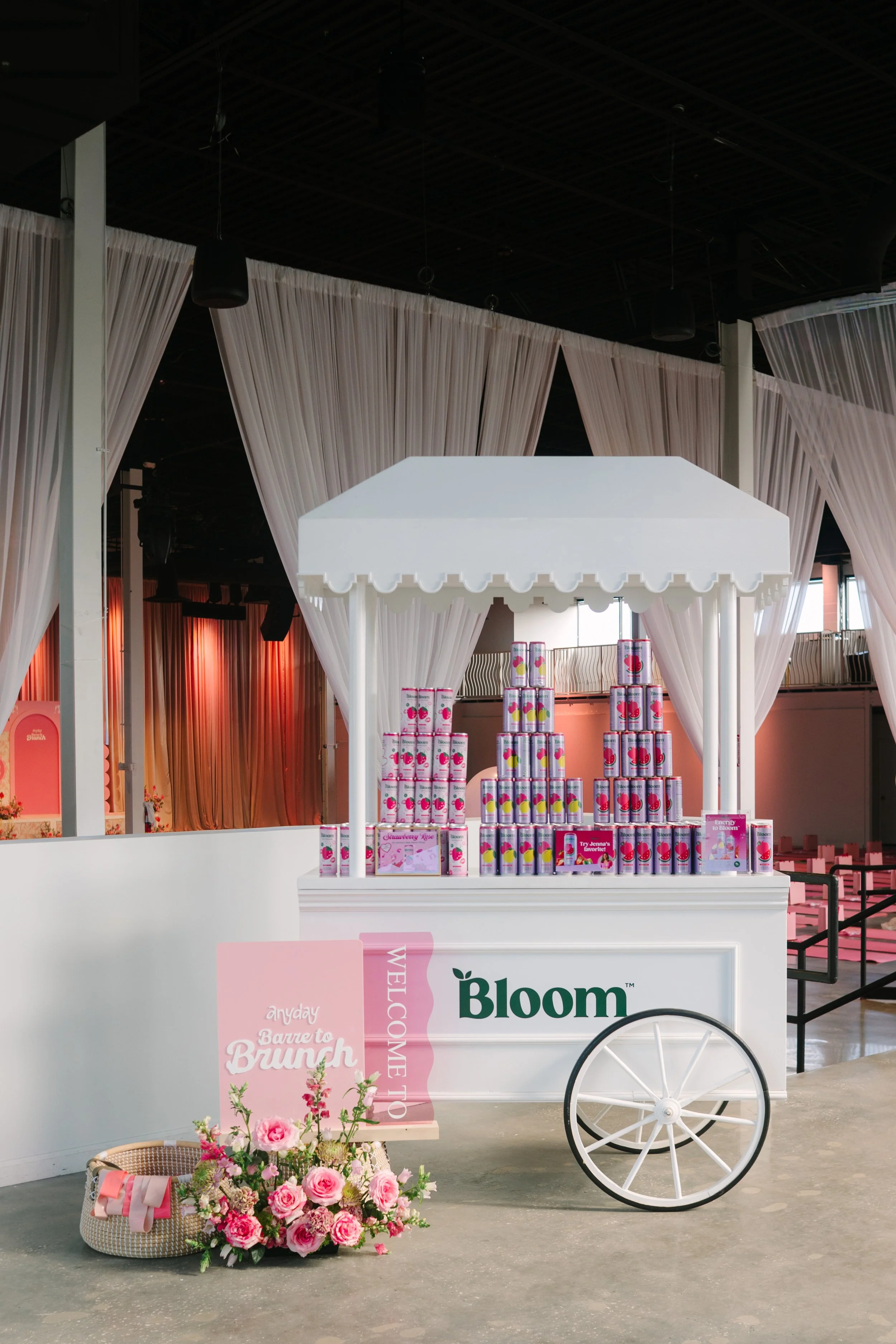 A Bloom flower and gift cart with a pink sign reading 'anyday Banchetto Brunch' surrounded by a pink flower bouquet and gift basket. Barre to Brunch