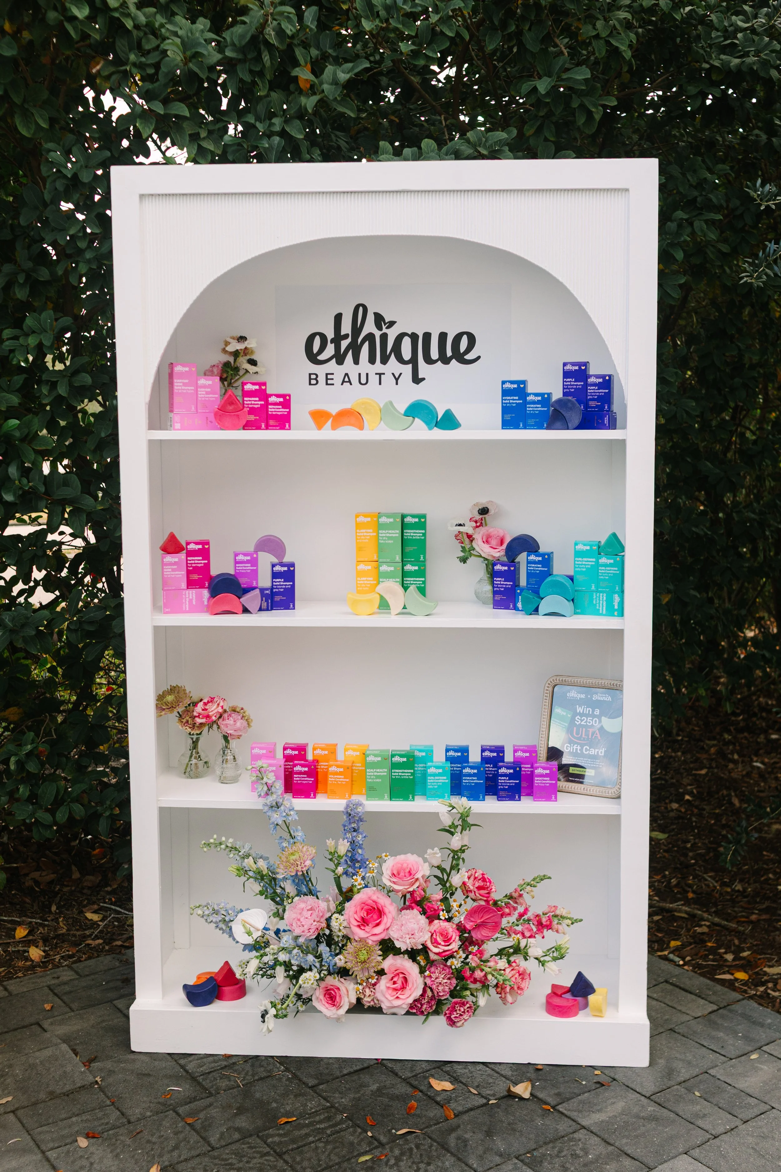  Ethique Beauty, arranged with pink, purple, blue, orange, green, and yellow packaging, decorated with pink and white flowers, set outdoors on a paved surface with green foliage in the background. Barre to Brunch with Jenna Palek