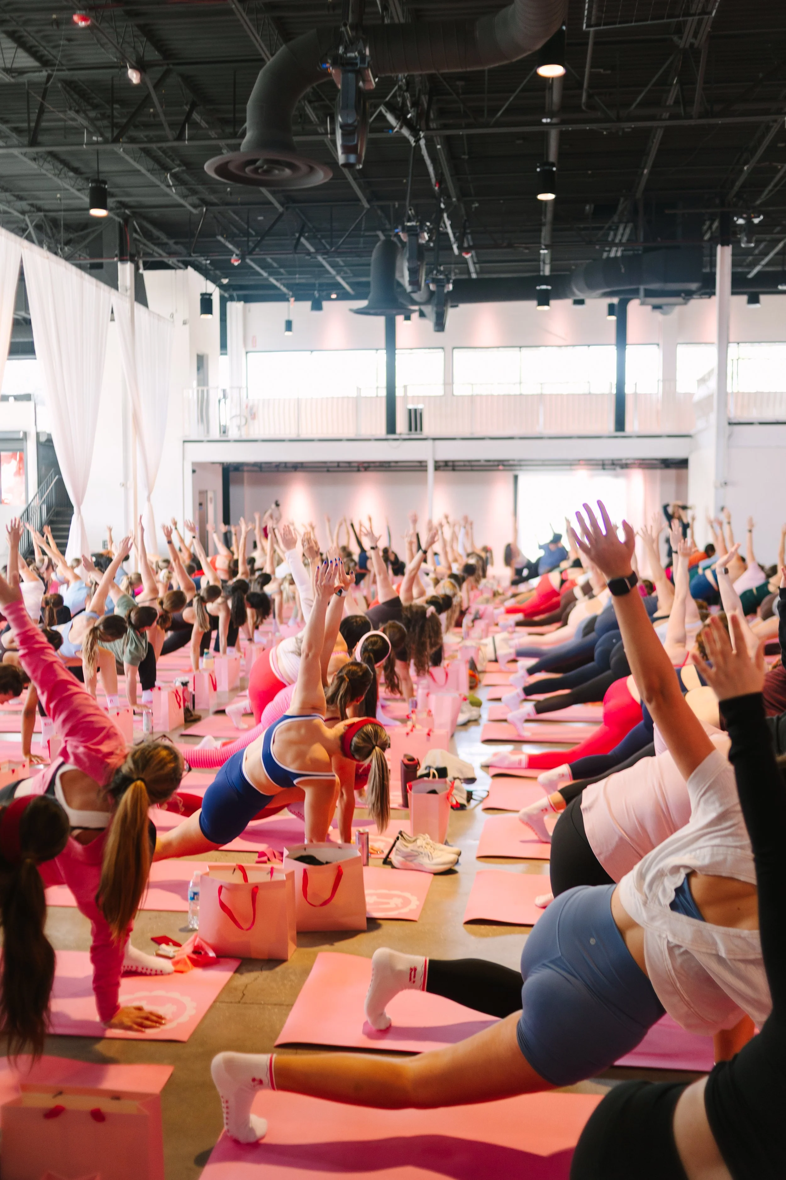 Group of women participating in a large, indoor yoga class, doing a side plank pose on pink mats with pink shopping bags beside each, in a bright, spacious studio. Barre to Brunch with Jenna Palek
