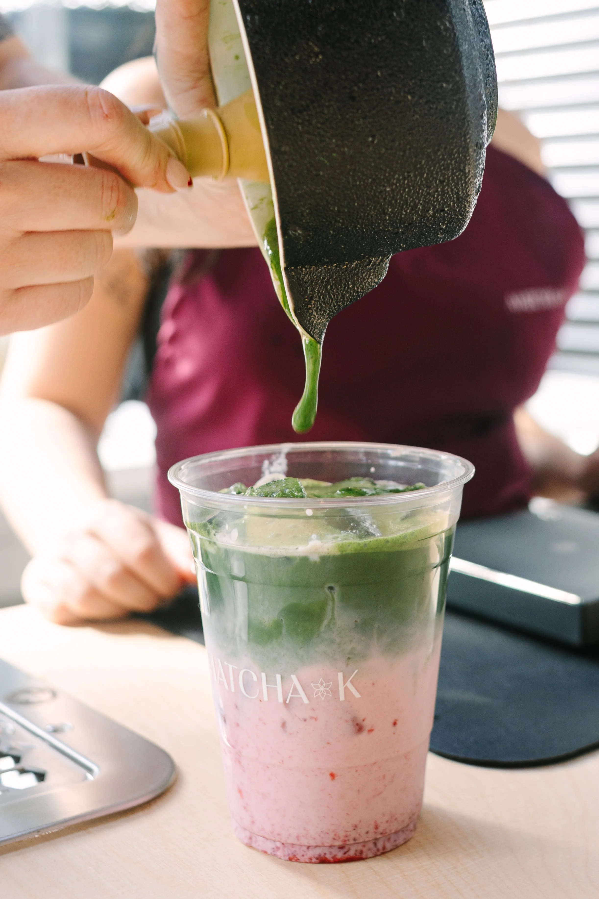 A person pouring green matcha latte into a clear plastic cup with a pink layered drink at the bottom. The cup is on a wooden table, and the scene is indoors with blinds in the background. Barre to Brunch with Jenna Palek with Sponsor Klassy