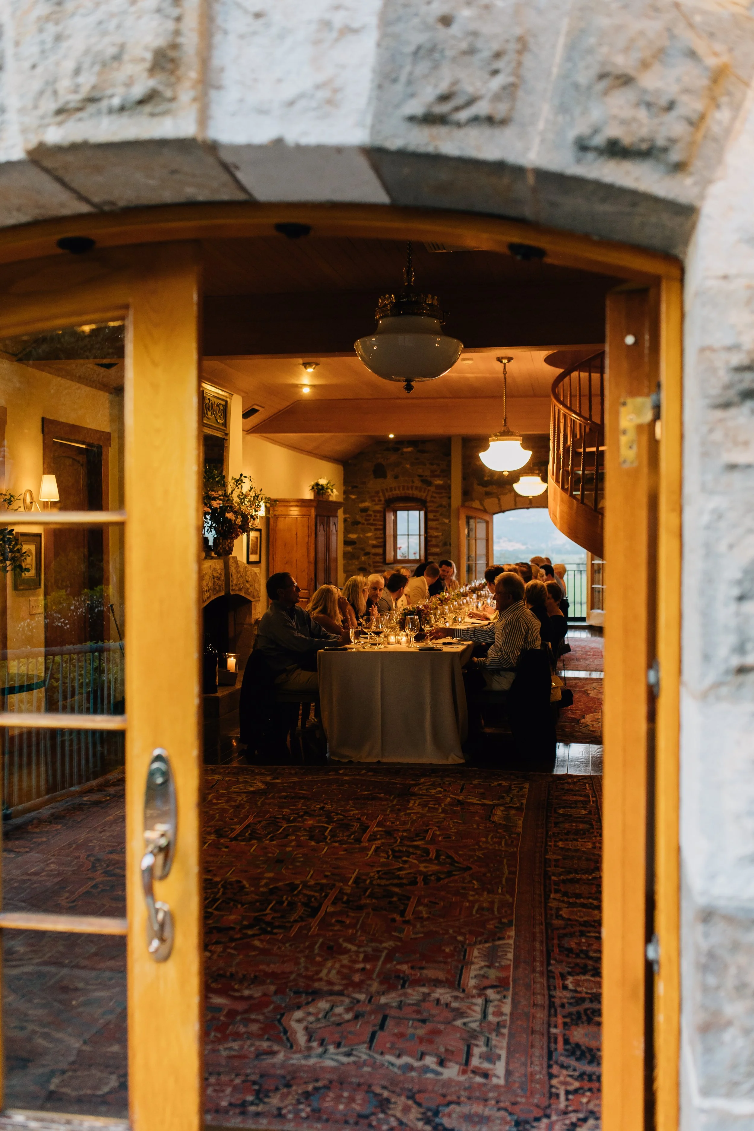 A view of a dinner gathering seen through a window with wooden framing, with people seated at a long table inside a cozy, rustic room with stone walls and wooden accents.