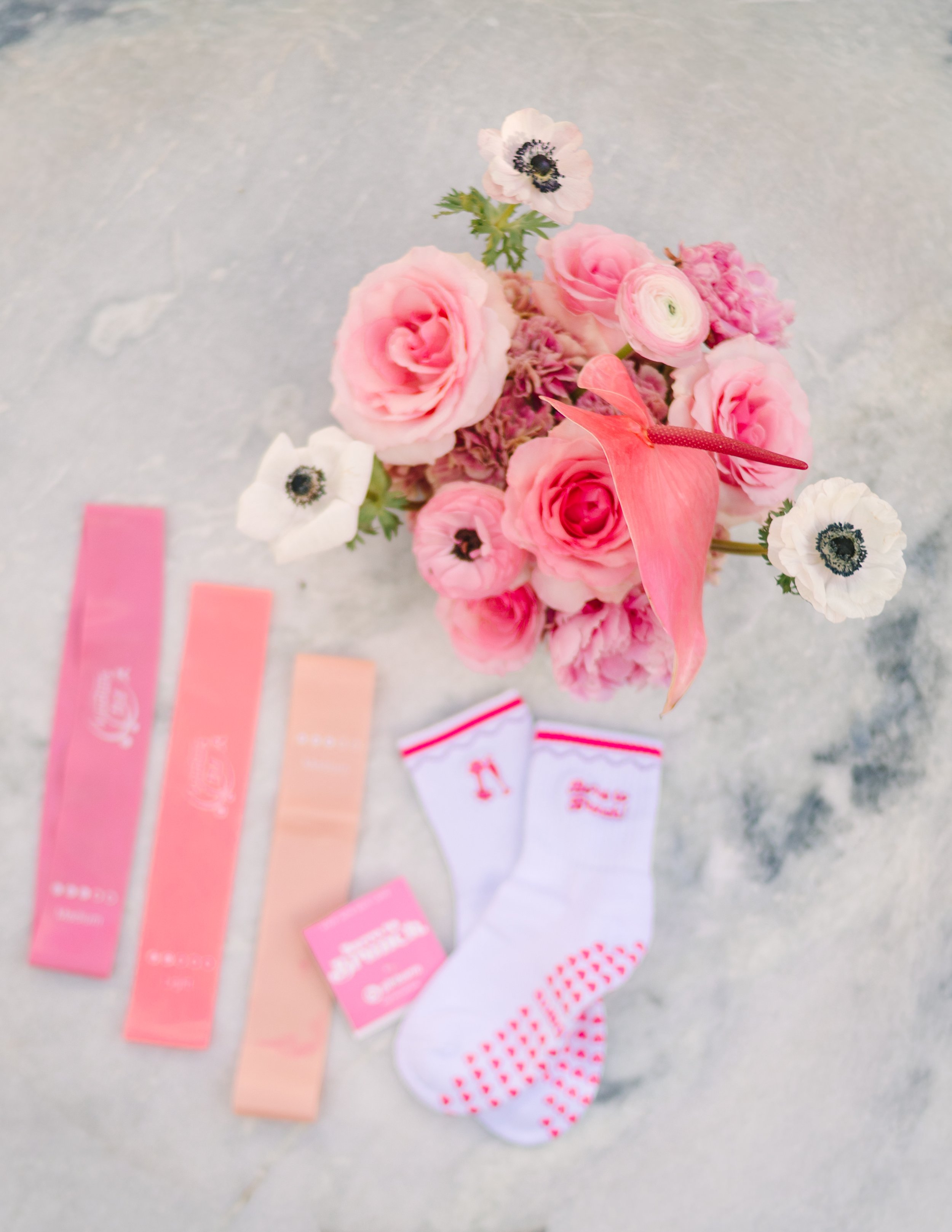 Pink and white flowers in a bouquet, with a pair of white socks with pink hearts, a small pink card, and pink ribbon bookmarks on a gray marble surface. Barre to Brunch with Jenna Palek