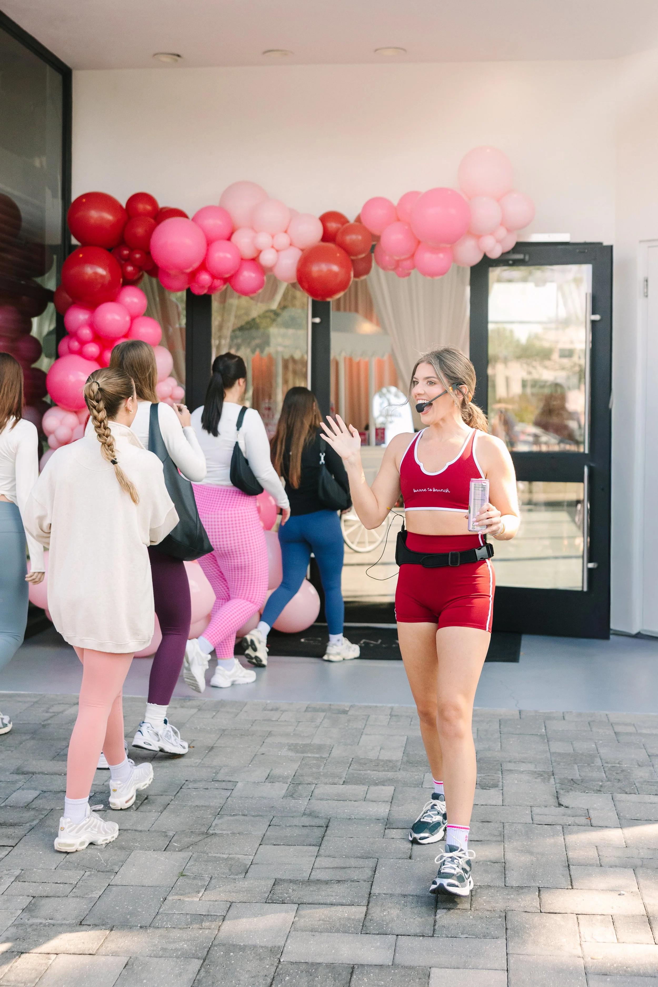 Indoor fitness class, instructor leading women in workout, pink and red balloon decorations outside glass door. Barre to Brunch with Jenna Palek