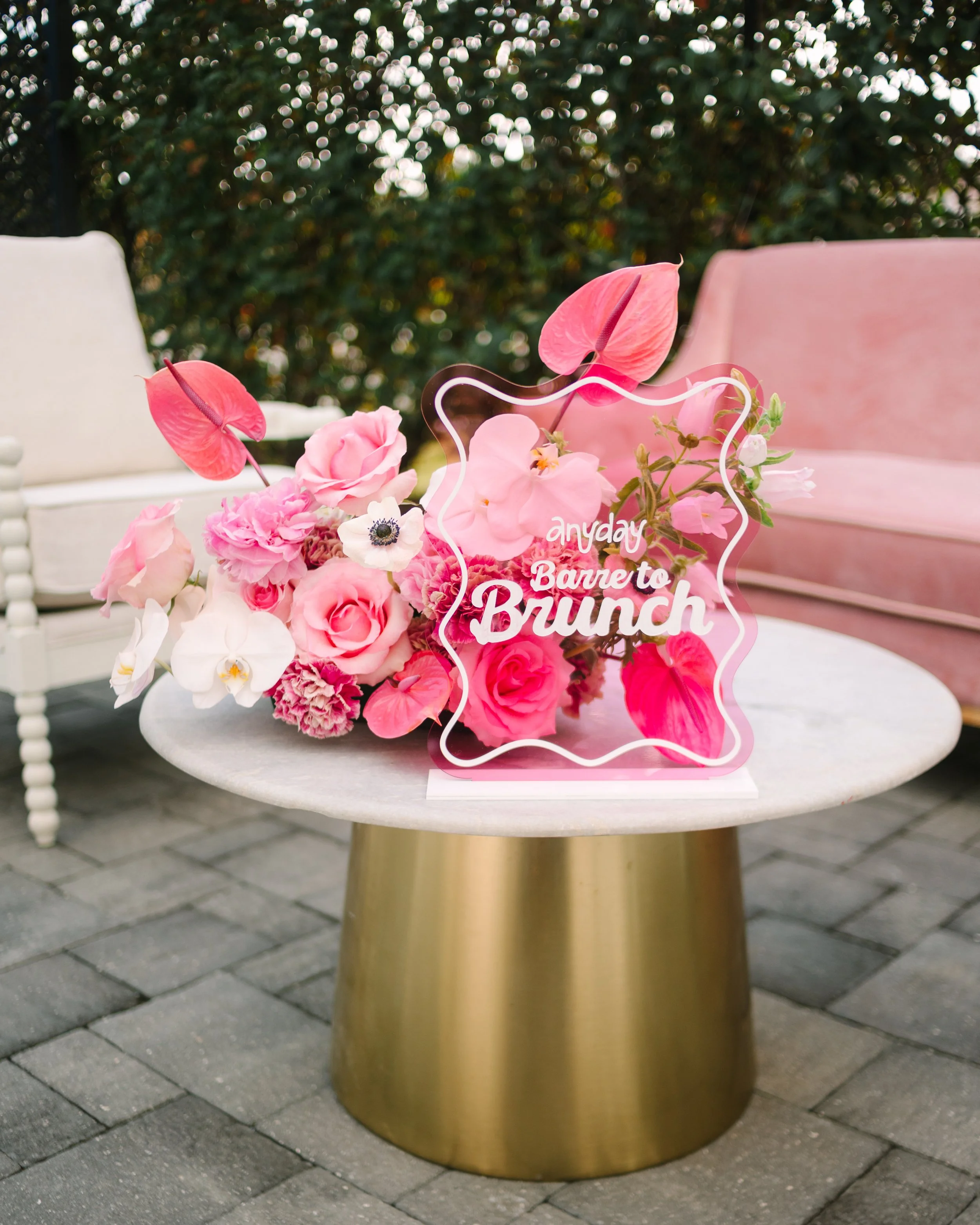 Pink and white floral bouquet with a neon sign that reads 'Anyday Brunch' on a white table with a pink sofa and white chairs in the background, outdoor setting with trees. Barre to Brunch with Jenna Palek