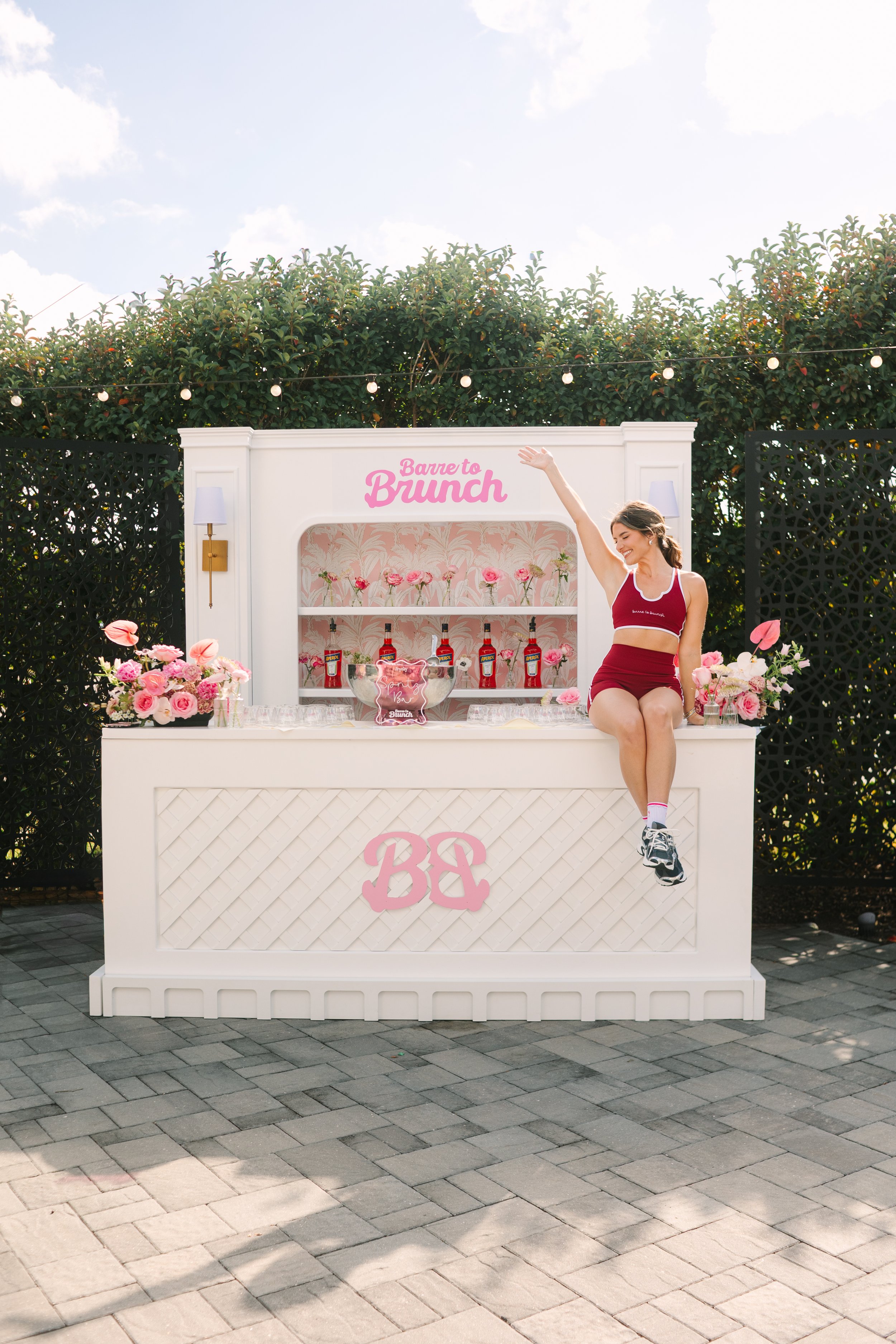 A woman sitting on a white bar with pink flowers and bottles, outdoor event with string lights, greenery, and a clear sky. Barre to Brunch with Jenna Palek