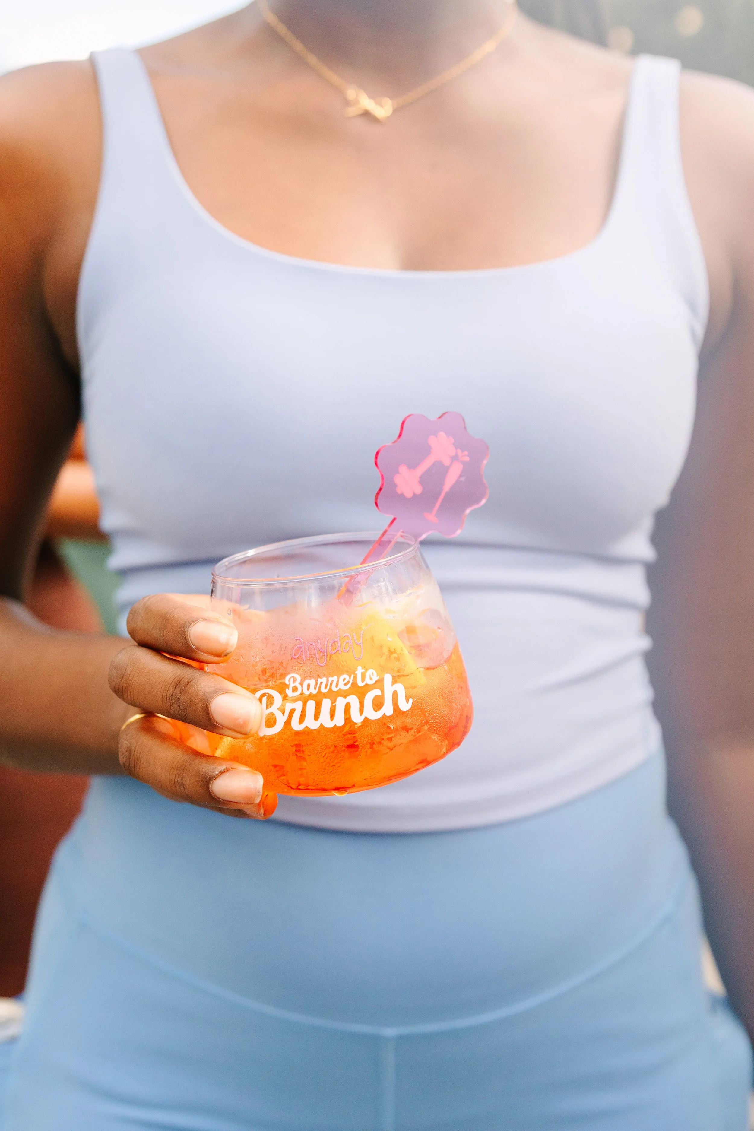 A woman in a white tank top holding a large glass with orange drink and ice, with a pink cake topper that has a number one and a flower, at an outdoor brunch event. Barre to Brunch with Jenna Palek