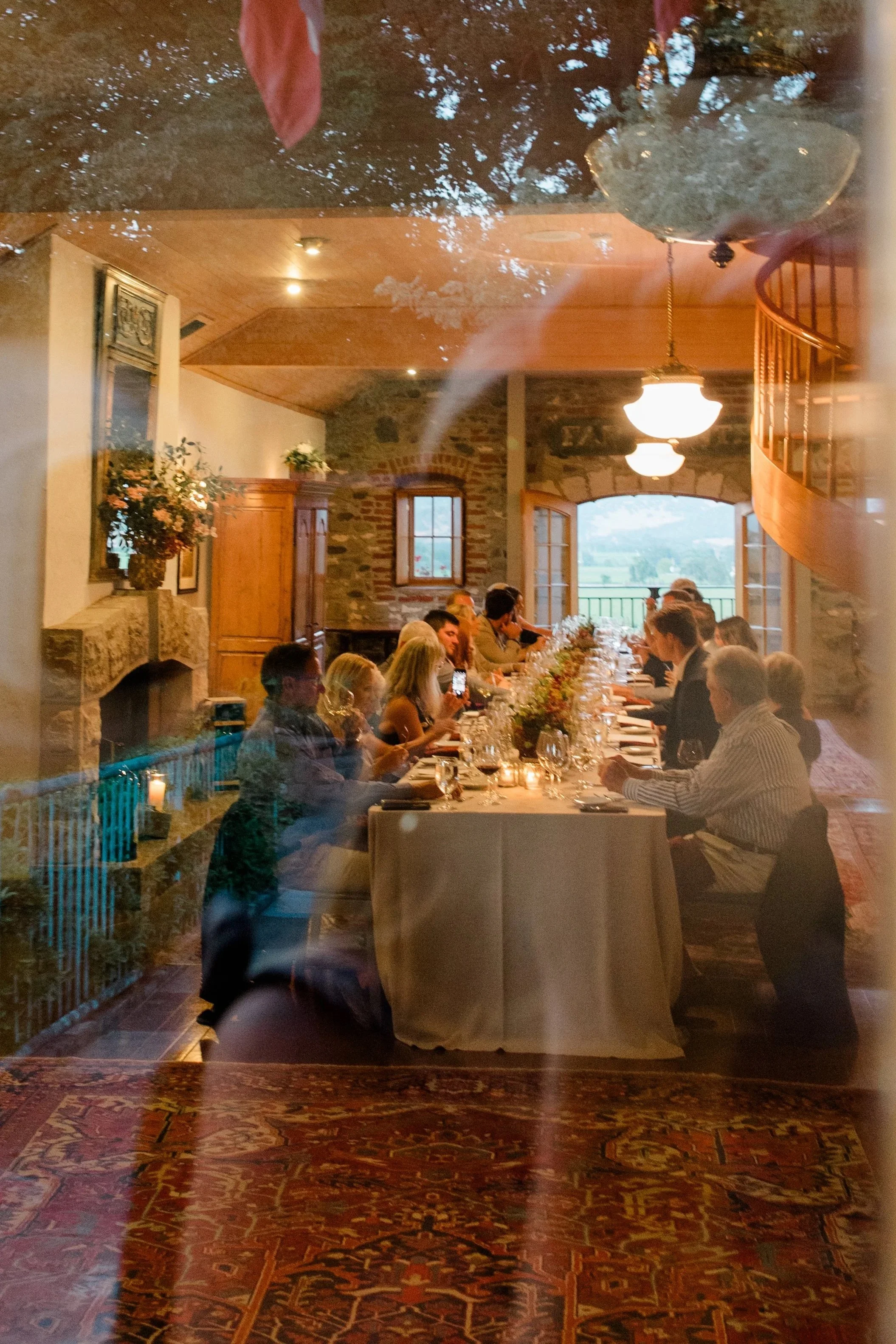 Reflected view of a group of people dining together at a long table in a cozy, rustic dining room with stone walls, wooden accents, and large windows showing outdoor scenery.