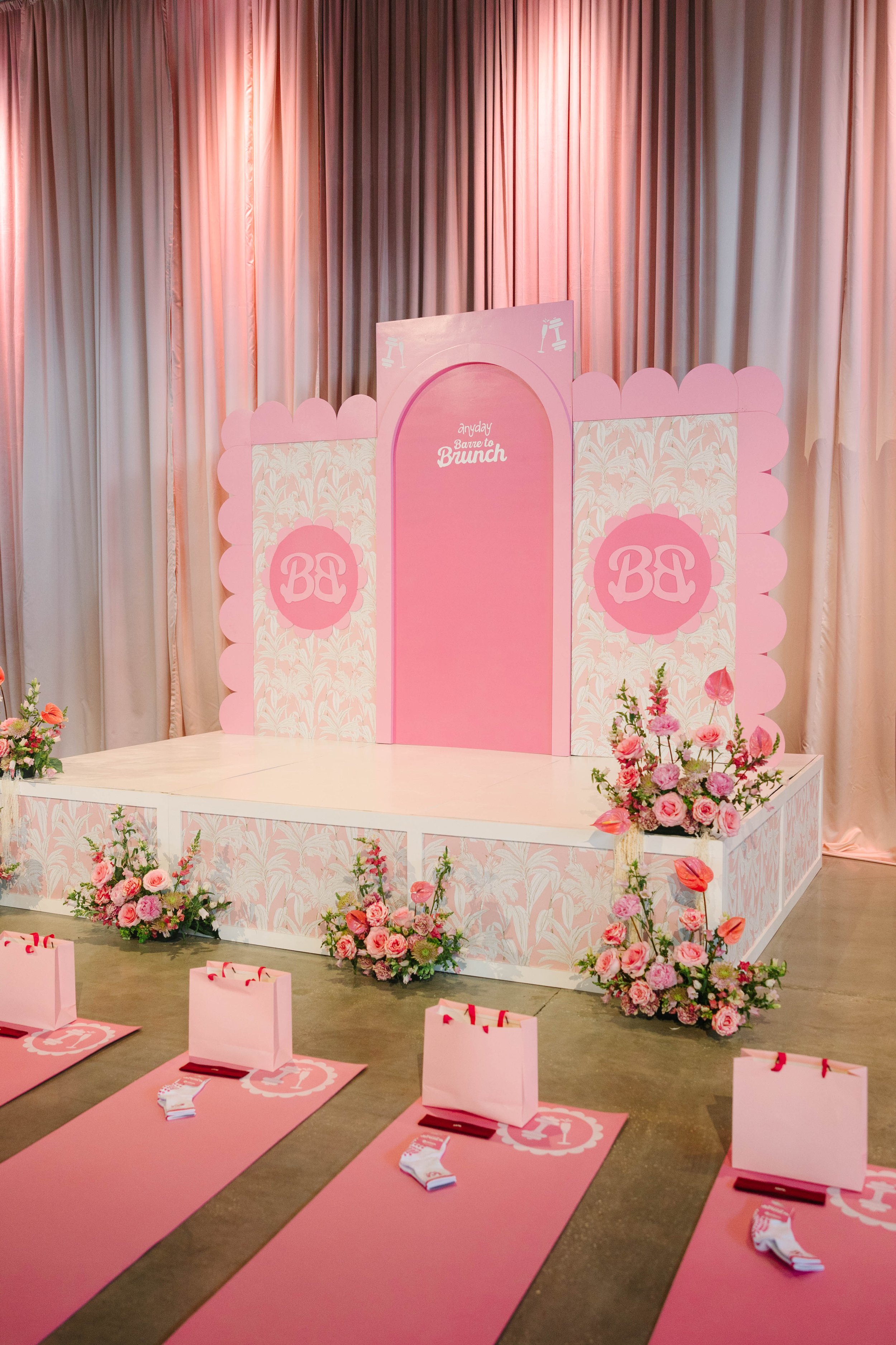 A pink-themed event stage decorated with flowers and a backdrop with the text 'Anyday Barre to Brunch' . There are pink mats with gift bags and socks arranged in front of the stage for a fitness or wellness event. Barre to Brunch Jenna Palek