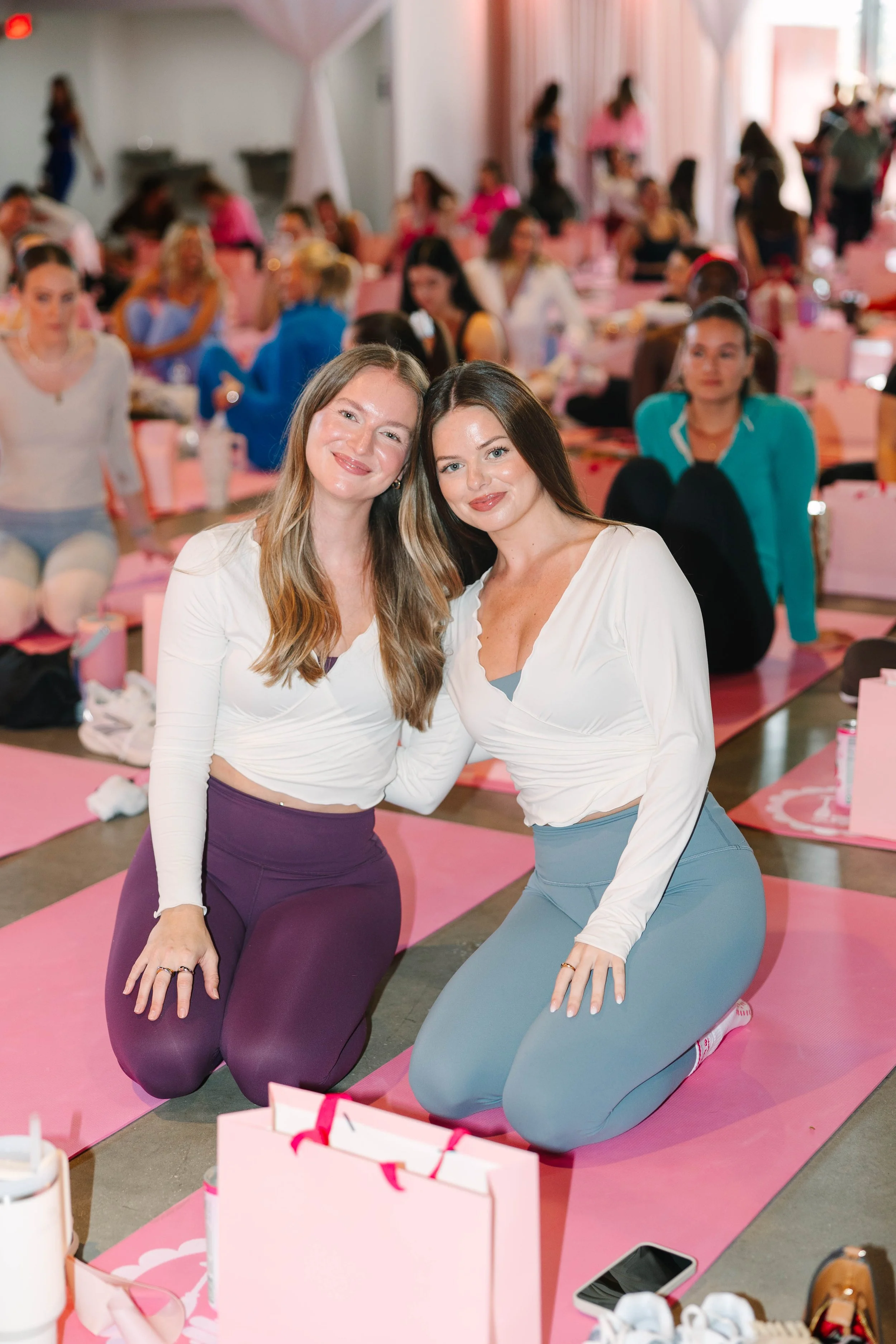 Two women in yoga pants and tops kneeling on pink mats amidst a group yoga class, smiling at the camera in a brightly lit studio. Barre to Brunch with Jenna Palek