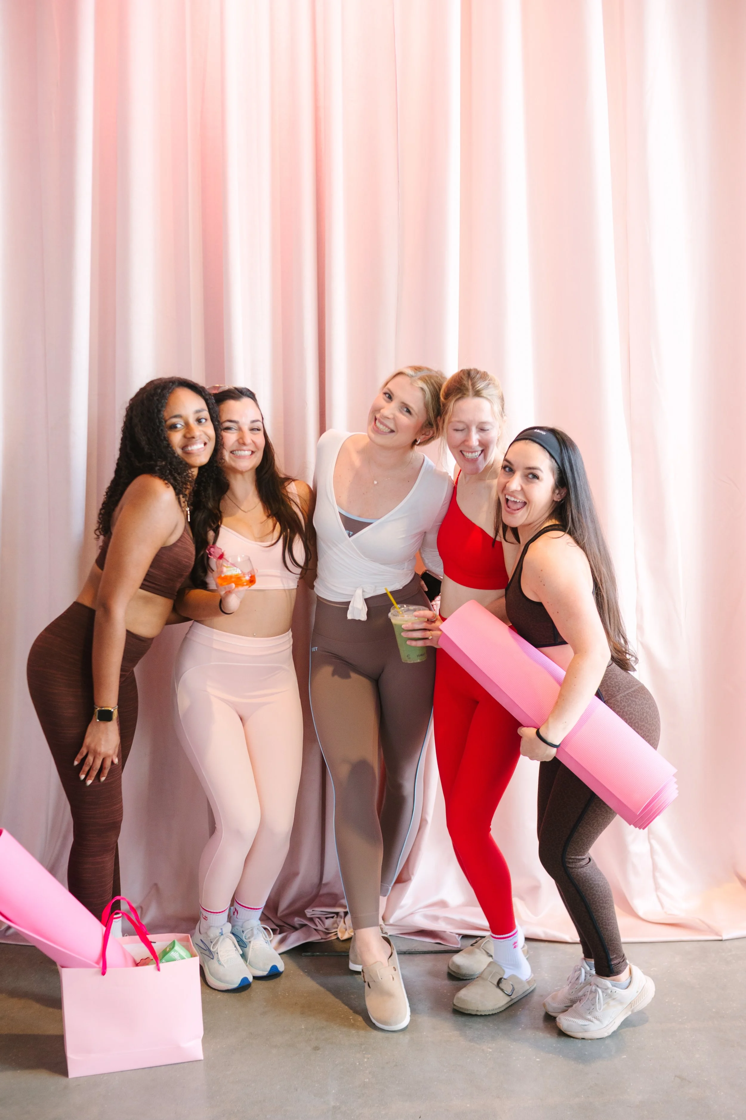 Group of five women in activewear smiling at a fitness or yoga class, holding drinks and yoga mats, in front of pink curtains.