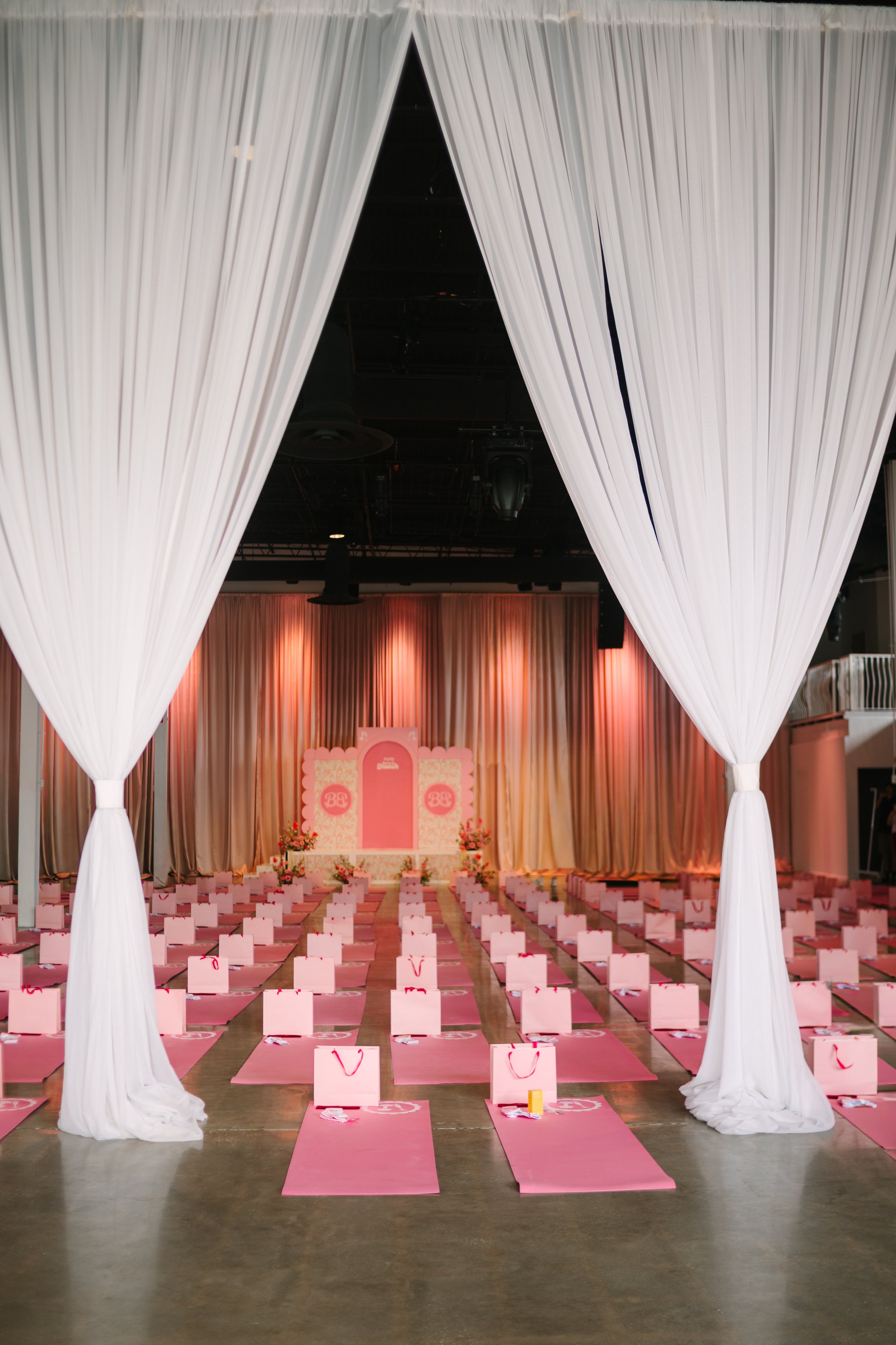 An indoor event space decorated in pink and white for a celebration or party, with an entrance draped, pink mats on the floor, and gift bags at each seat, leading to a stage with curtains and floral arrangements. Barre to Brunch with Jenna Palek 