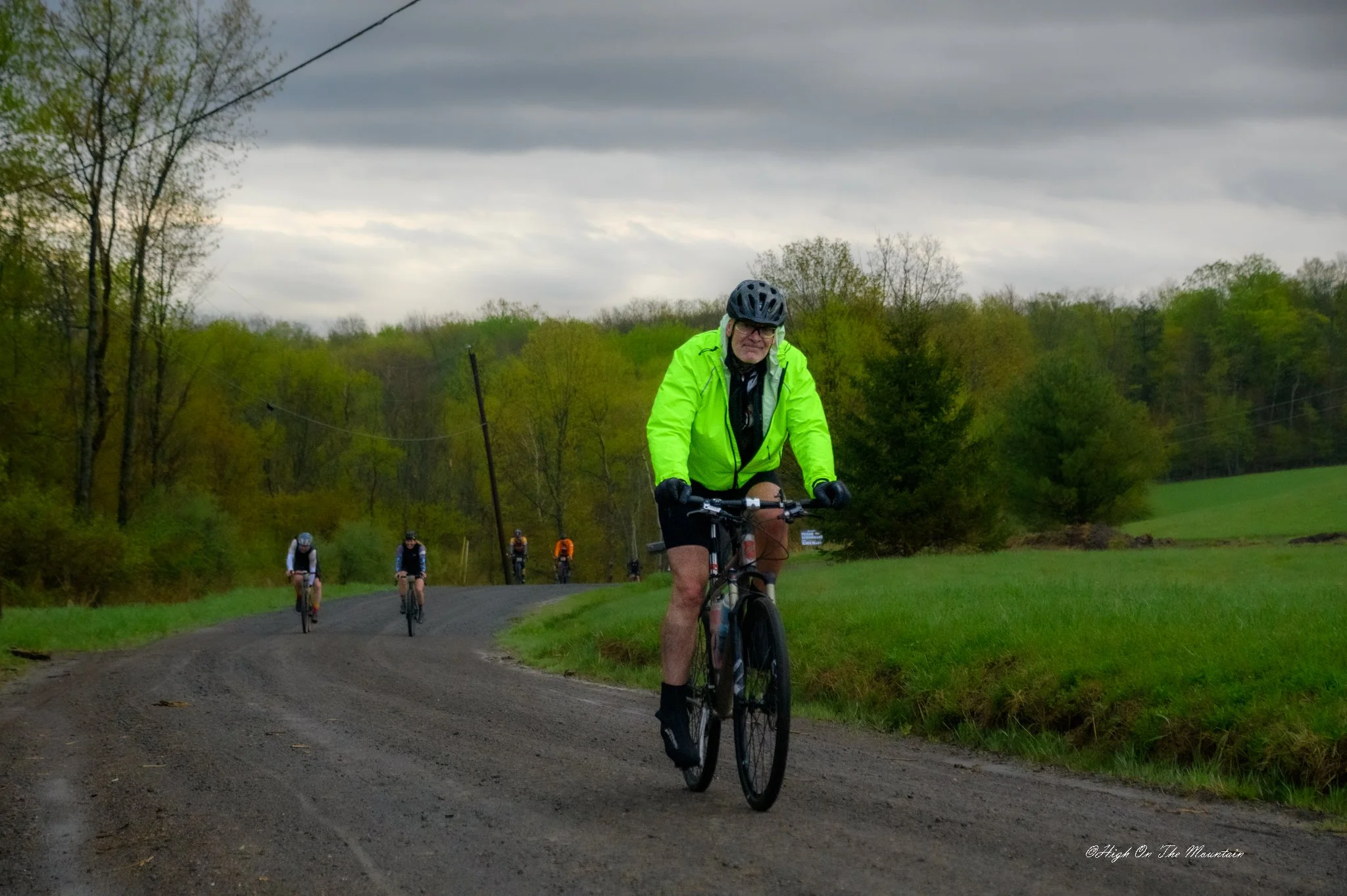 A group of people riding bicycles on a dirt path through a green countryside landscape under a cloudy sky. The lead cyclist is wearing a bright yellow jacket and black helmet.