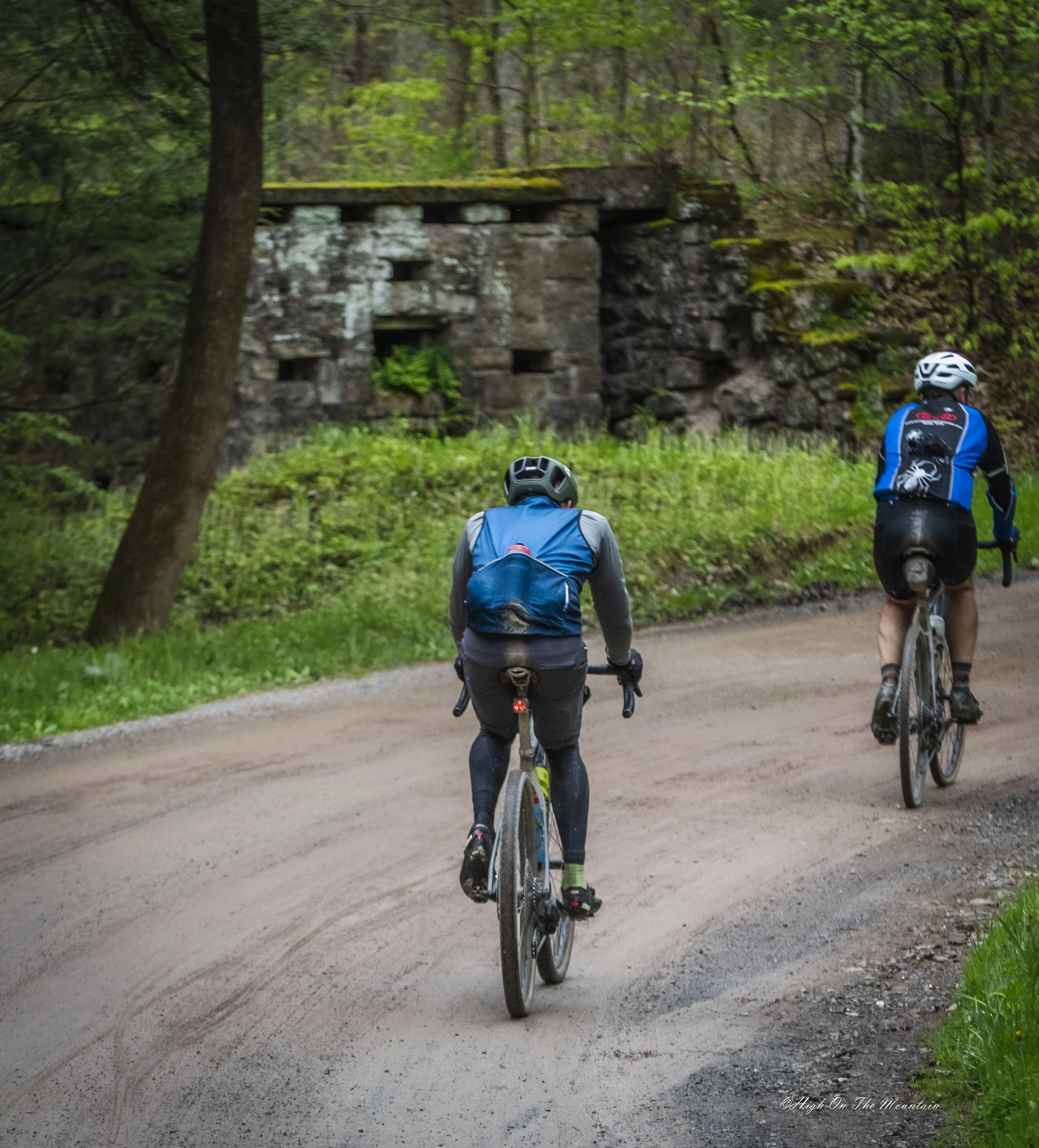 Two cyclists riding on a dirt forest trail surrounded by green trees, with an old stone structure in the background.