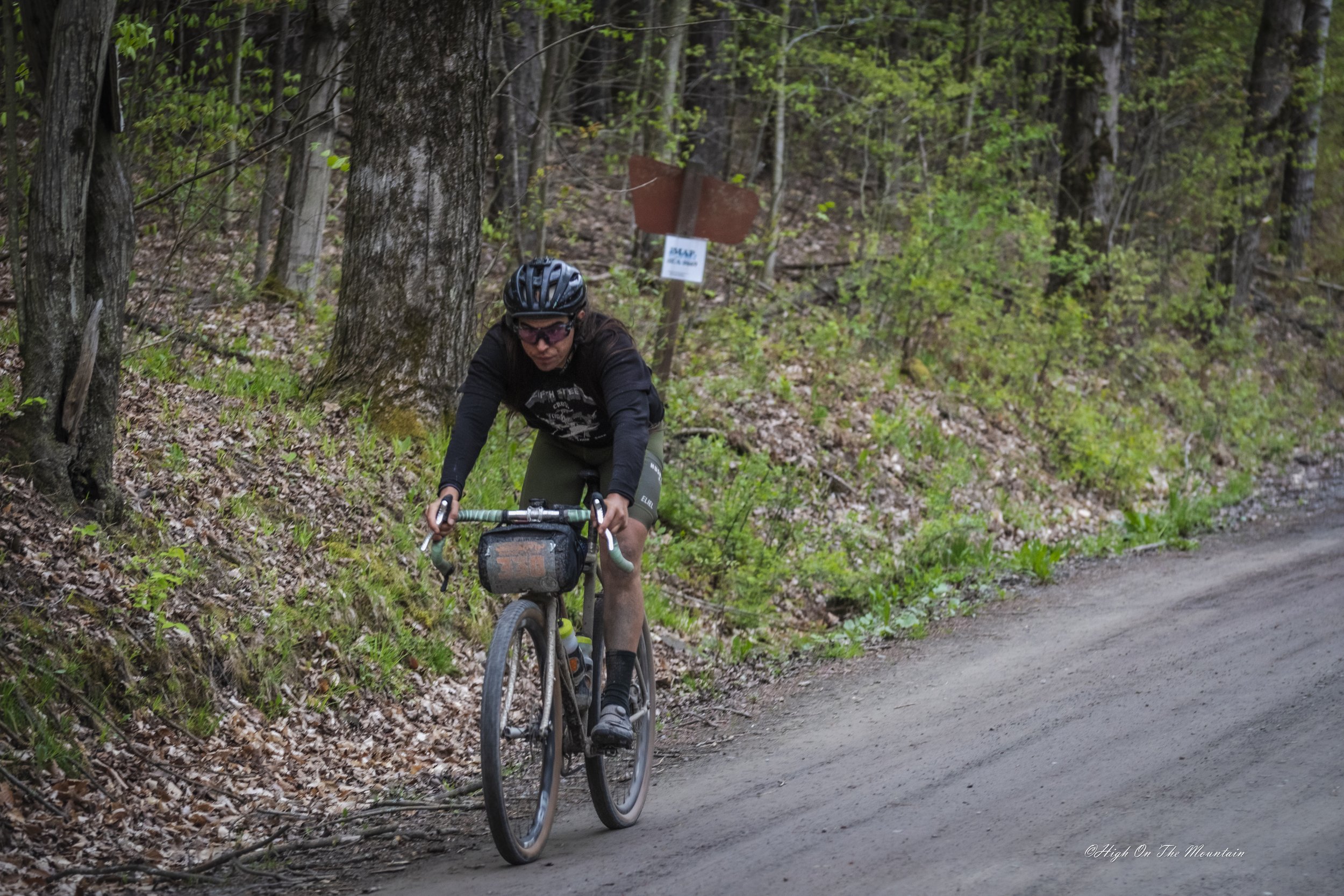 A person mountain biking on a trail through a wooded area with trees and green leaves.
