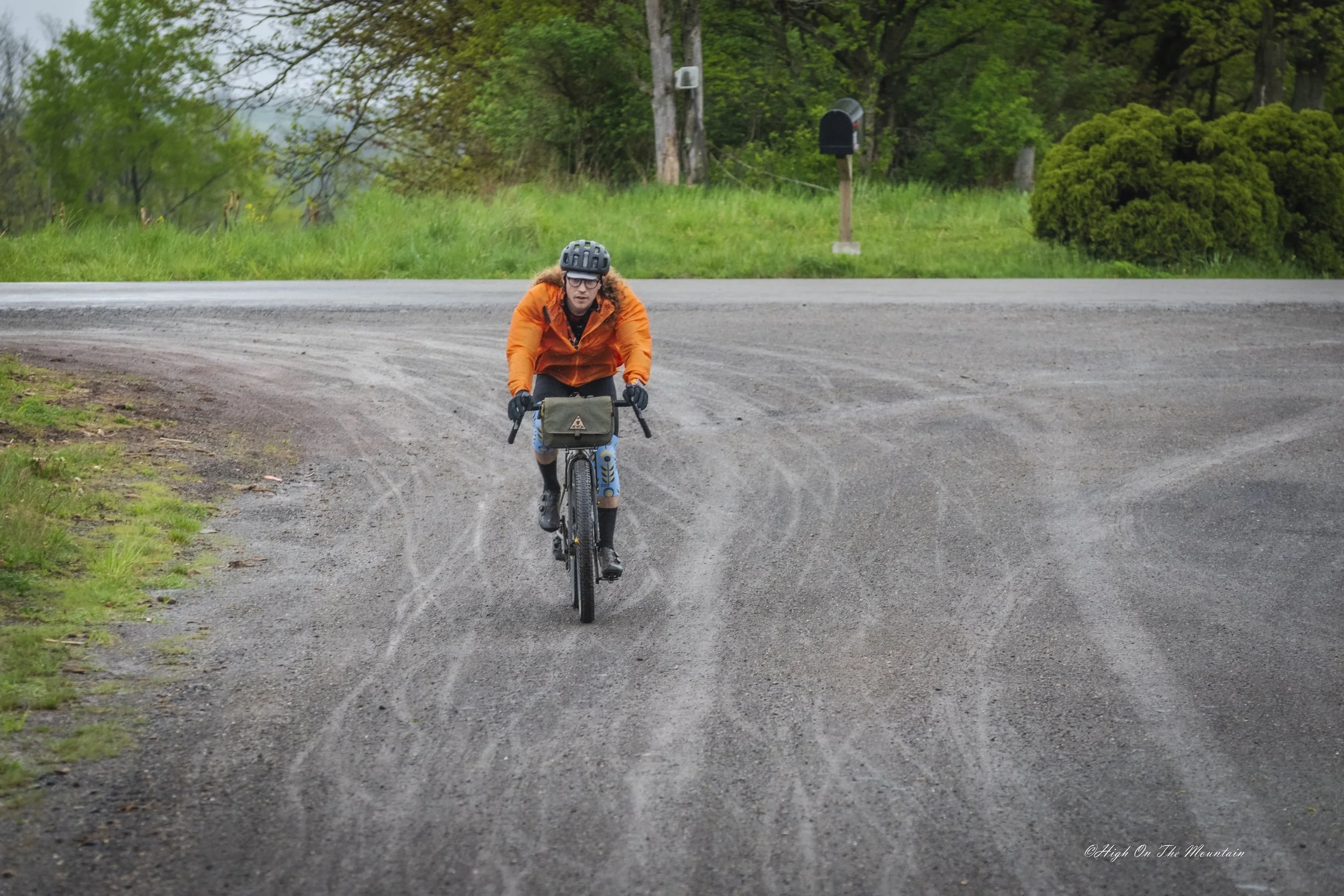 Person riding a mountain bike on a dirt road in a green forested area, wearing an orange jacket and a black helmet.