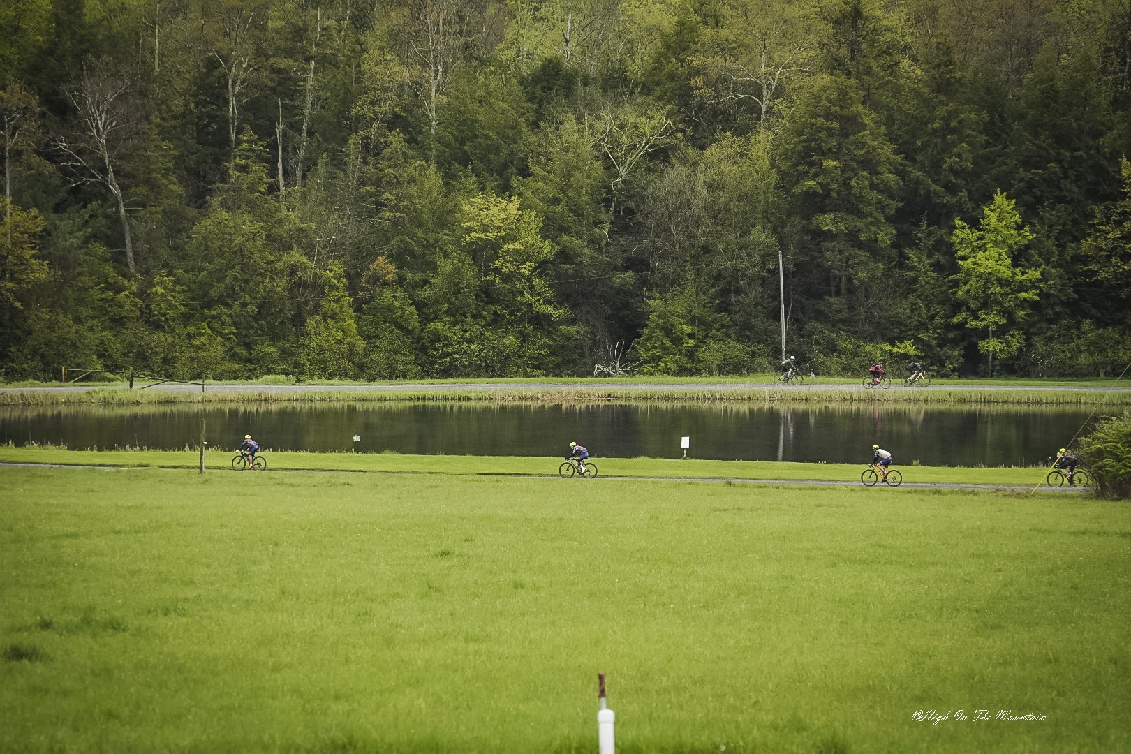 Bicyclists riding on a path beside a lake with a dense forest in the background.
