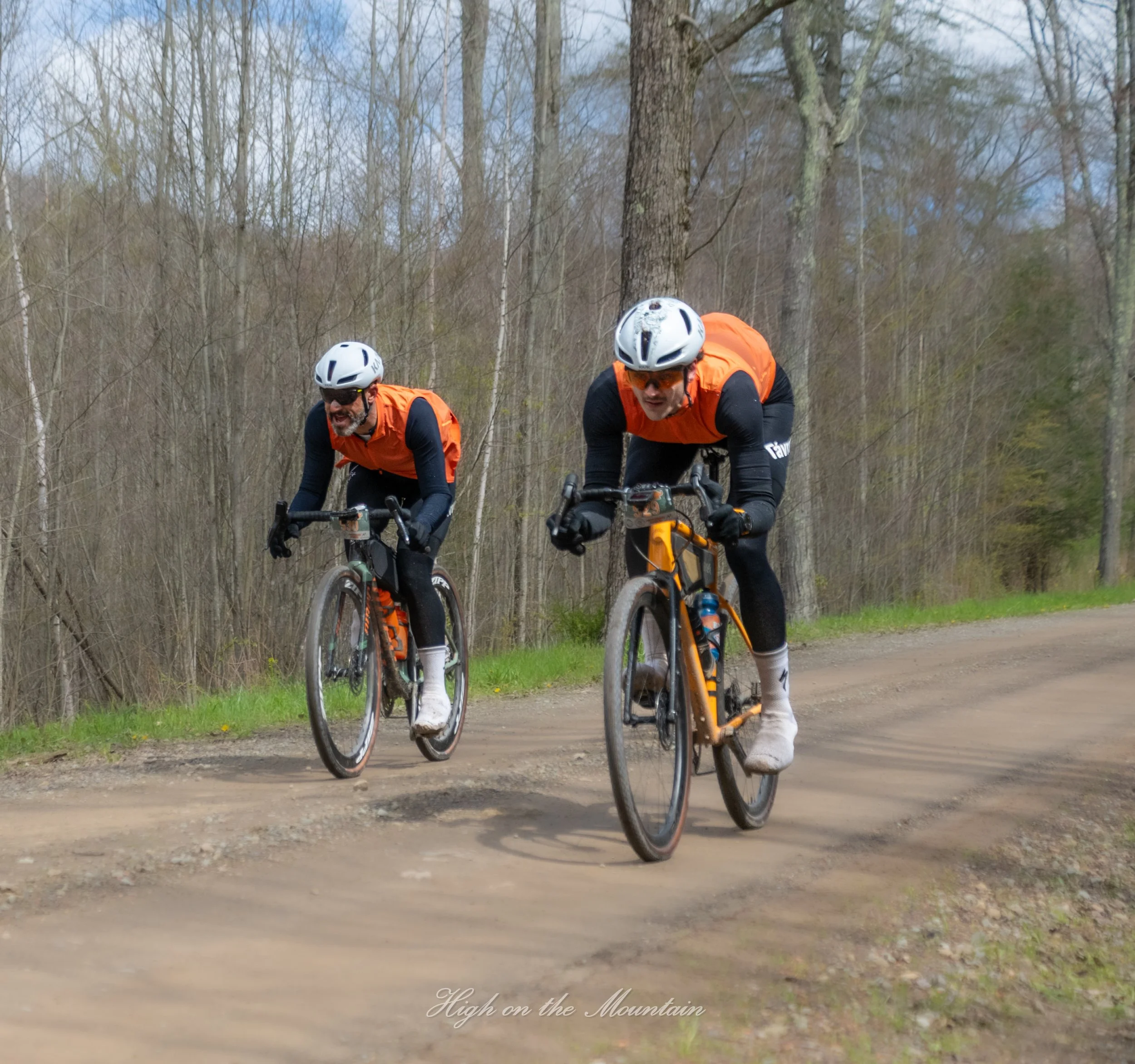 Two men wearing orange vests and white helmets riding mountain bikes through a wooded trail.