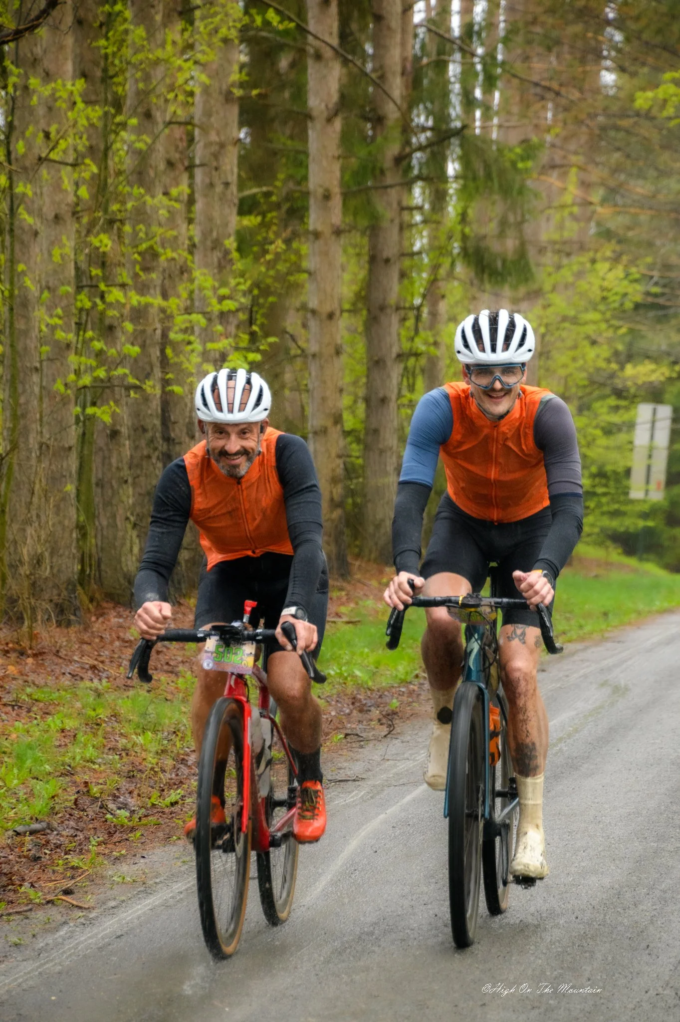 Two men riding bikes on a forest trail, wearing helmets and orange vests, smiling and enjoying the ride through green trees.