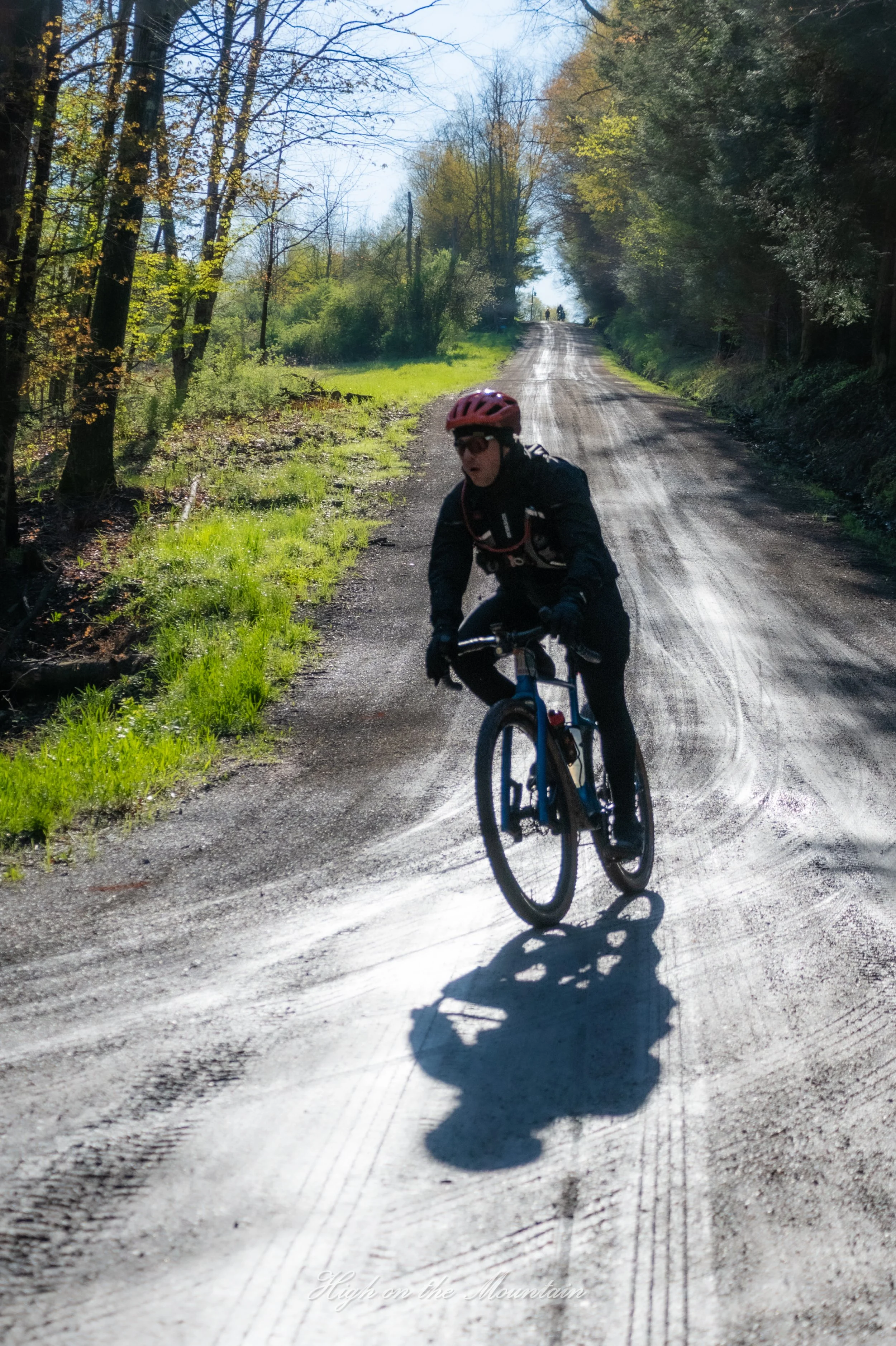 A person in black cycling gear, red helmet, and sunglasses riding a mountain bike on a dirt trail through a forest. The person appears to be racing or enjoying an outdoor ride, with several people visible in the distance on the tree-lined trail. Sunl