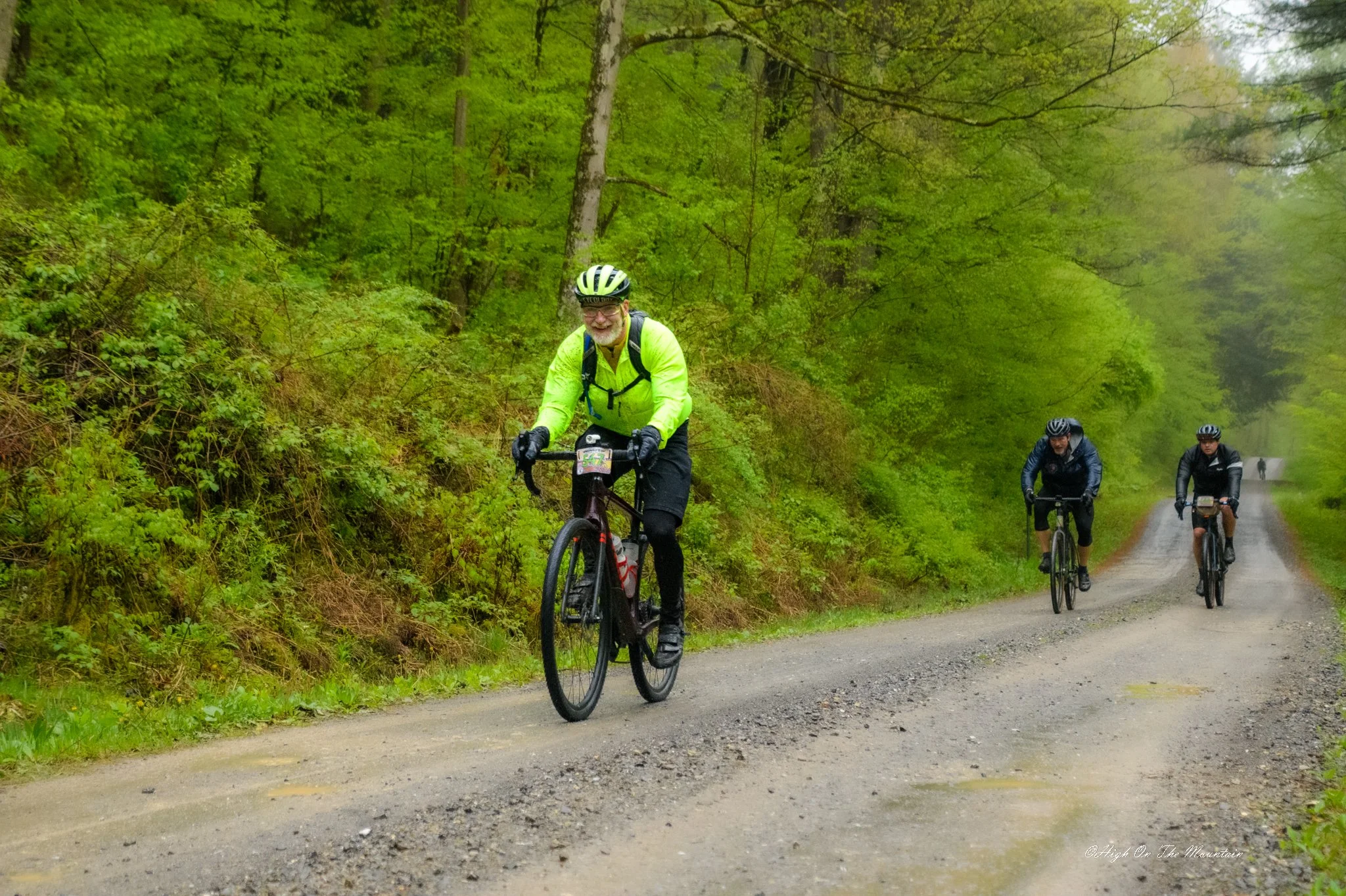 Three cyclists riding on a gravel trail through a lush green forest, with trees and bushes on either side.