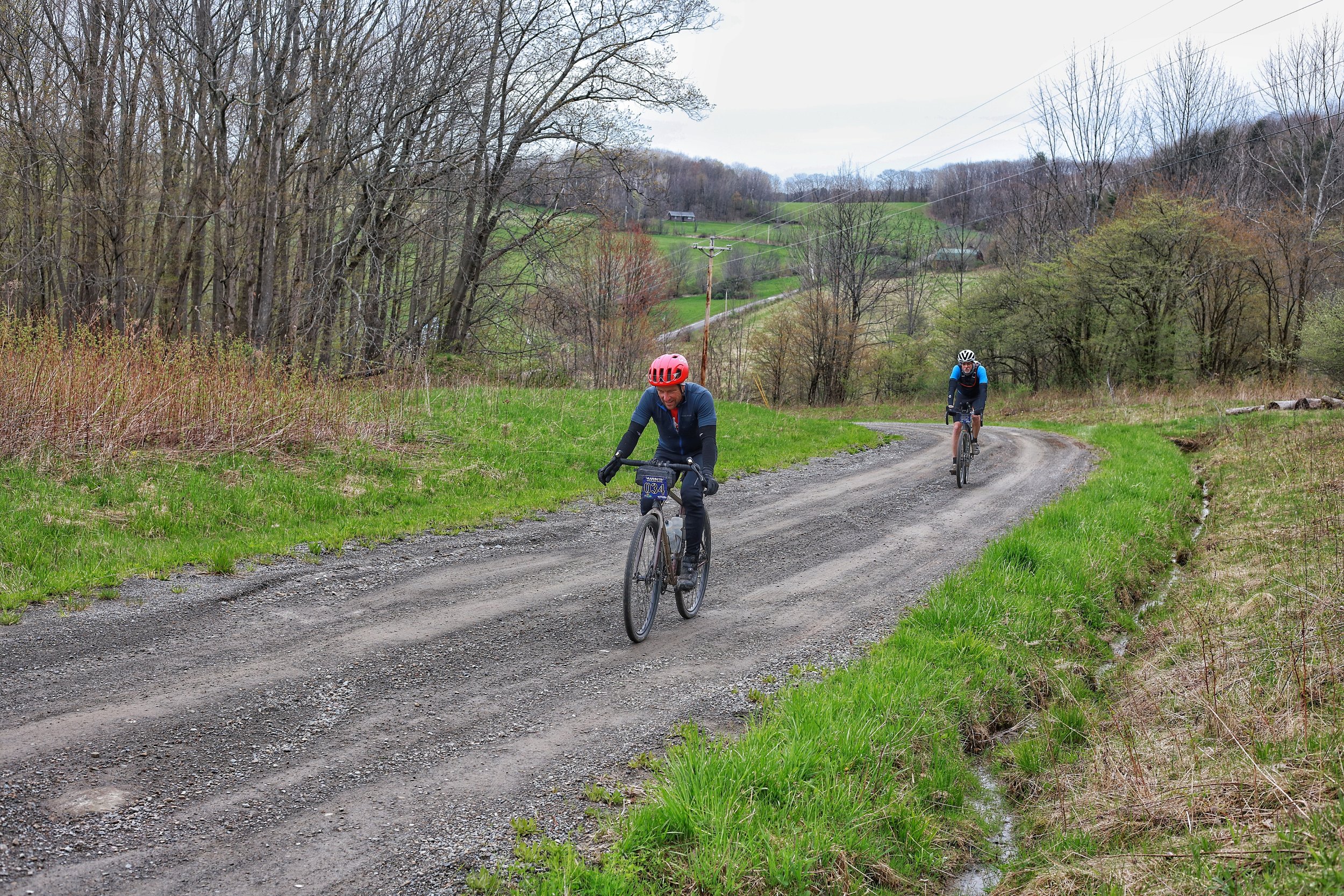 Two cyclists riding on a dirt road in a rural landscape with green grass, trees, and rolling hills in the background.