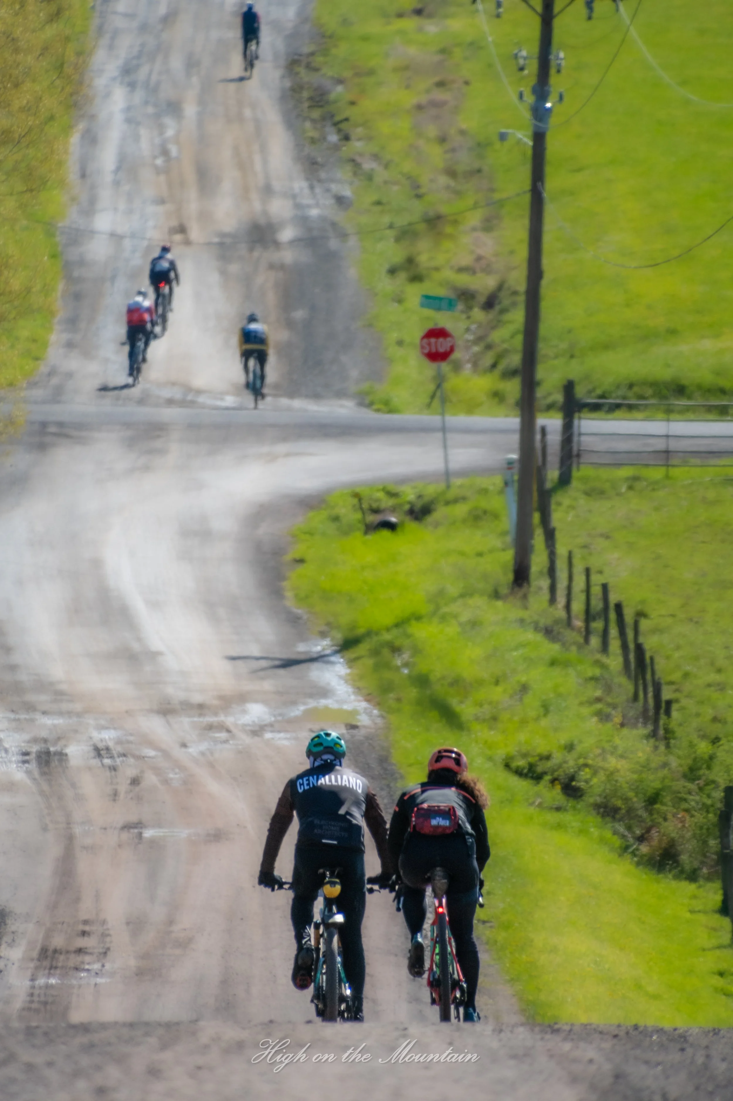 Bikers riding on a dirt mountain trail with green grass on the sides and a stop sign visible on the right side of the trail.