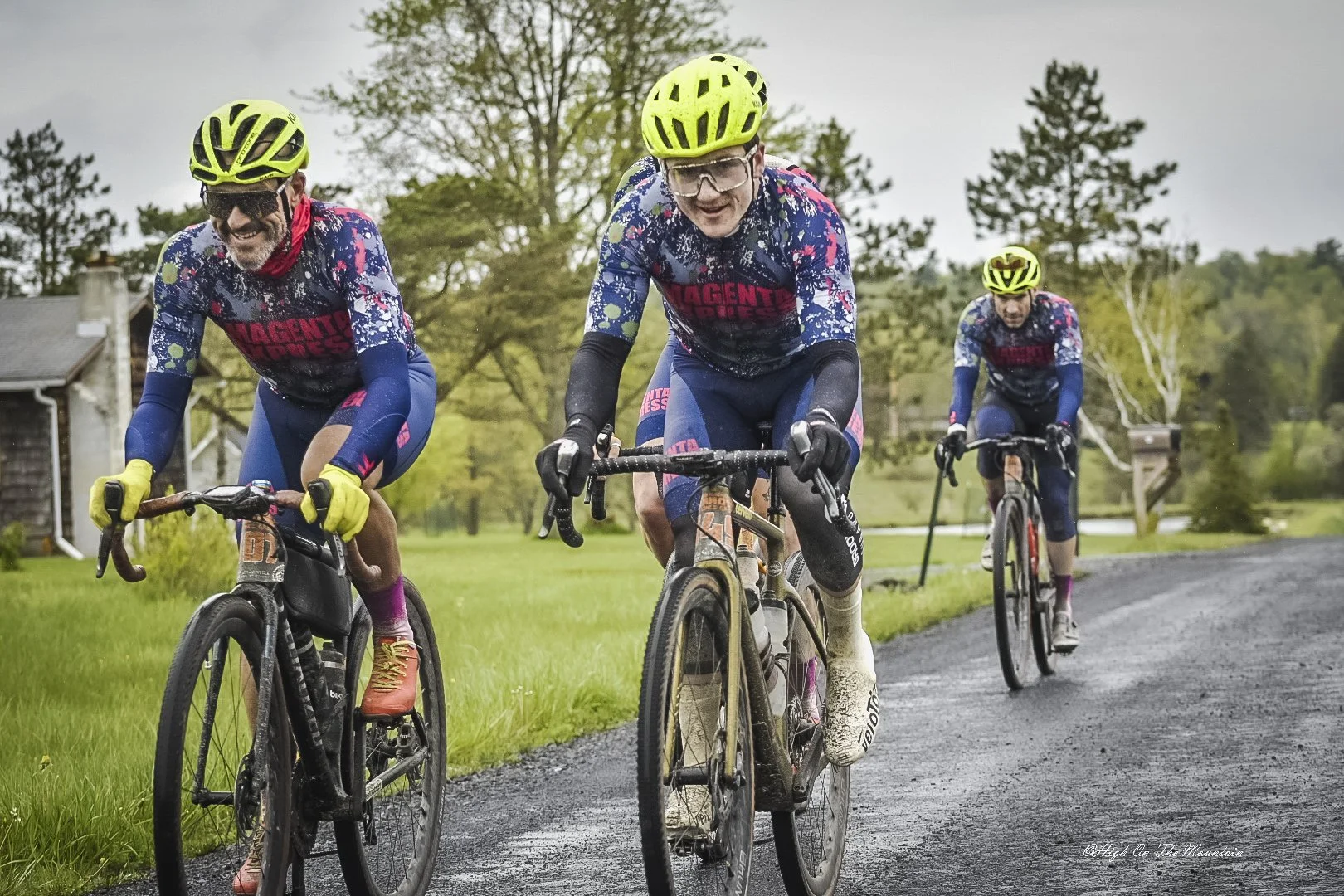 Three cyclists riding on a gravel path in a rural area with trees and a house in the background, wearing colorful cycling gear and yellow helmets during overcast weather.