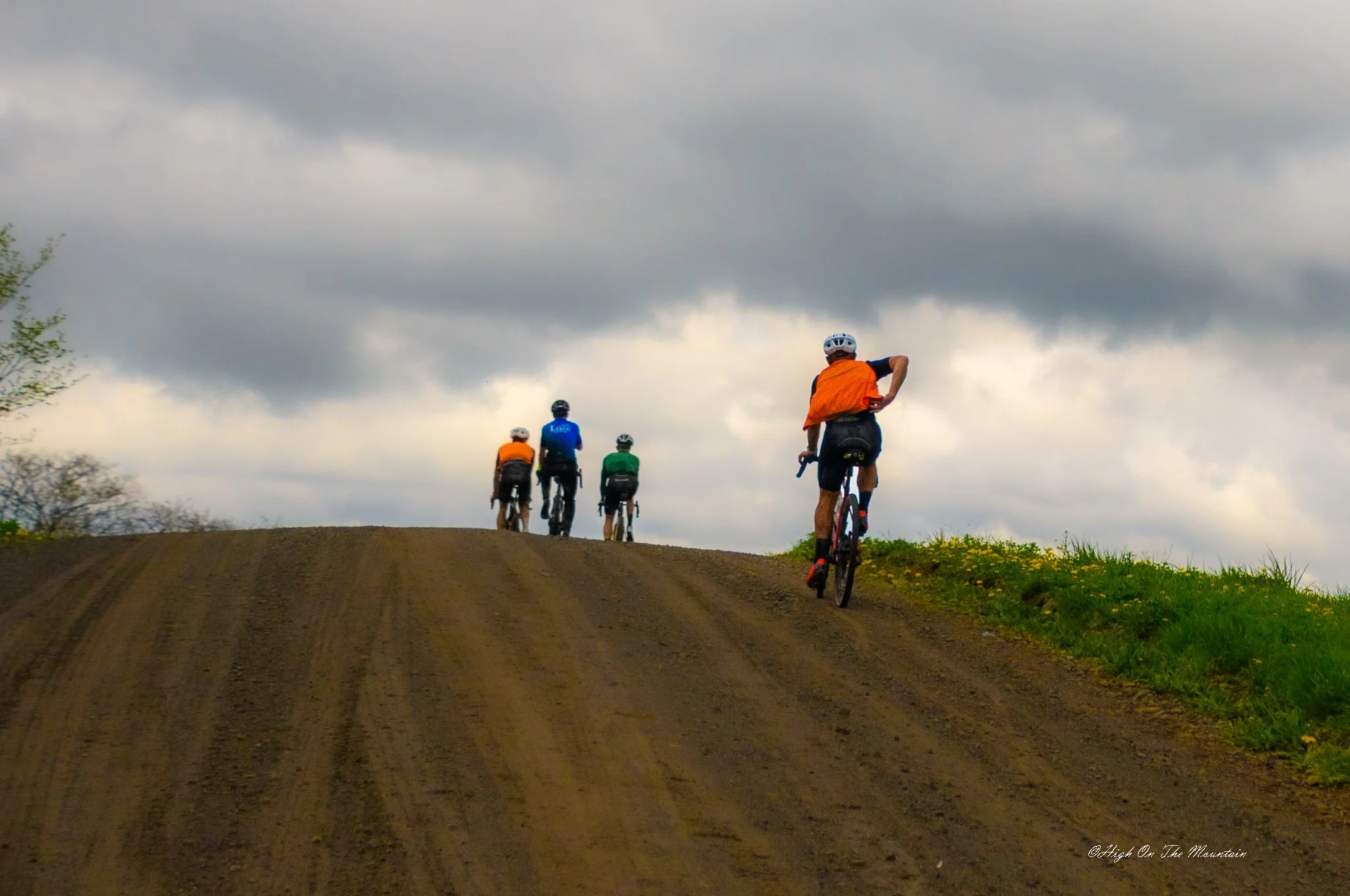 Four cyclists riding on a dirt path on a cloudy day, with one cyclist in the foreground wearing an orange vest, and three others further ahead in blue, green, and orange jerseys.