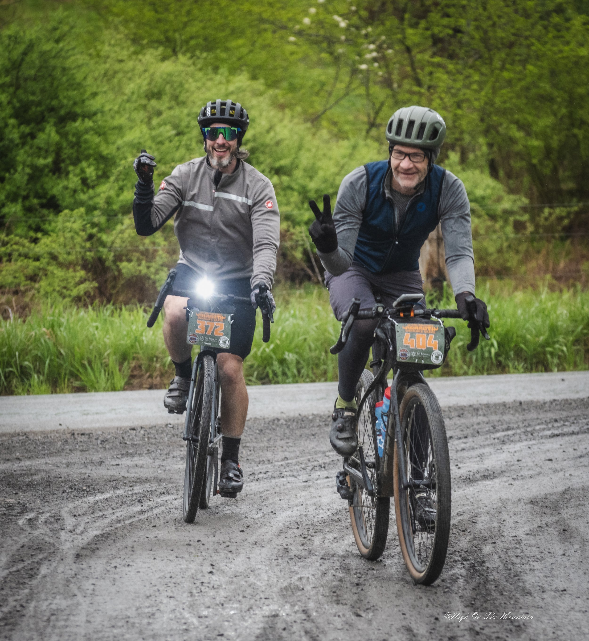 Two men riding mountain bikes on a dirt path in a lush green forest. The man on the left is smiling, wearing a gray jacket, black gloves, sunglasses, and a black helmet. The man on the right is smiling, making a peace sign with his right hand, wearin