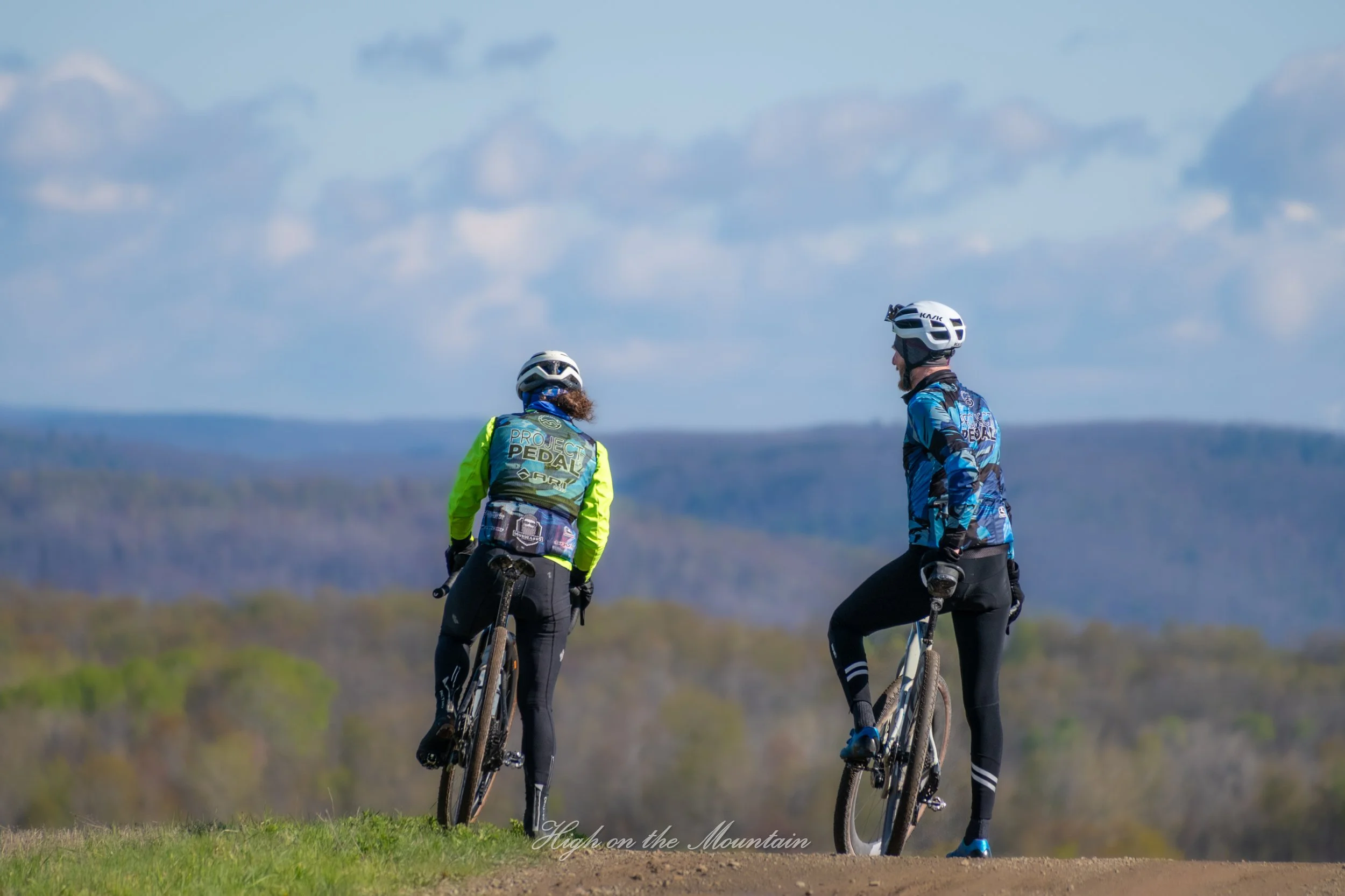 Two mountain bikers with helmets and sports gear standing with their bikes outdoors on a grassy trail, overlooking a distant landscape with hills and trees under a partly cloudy sky.