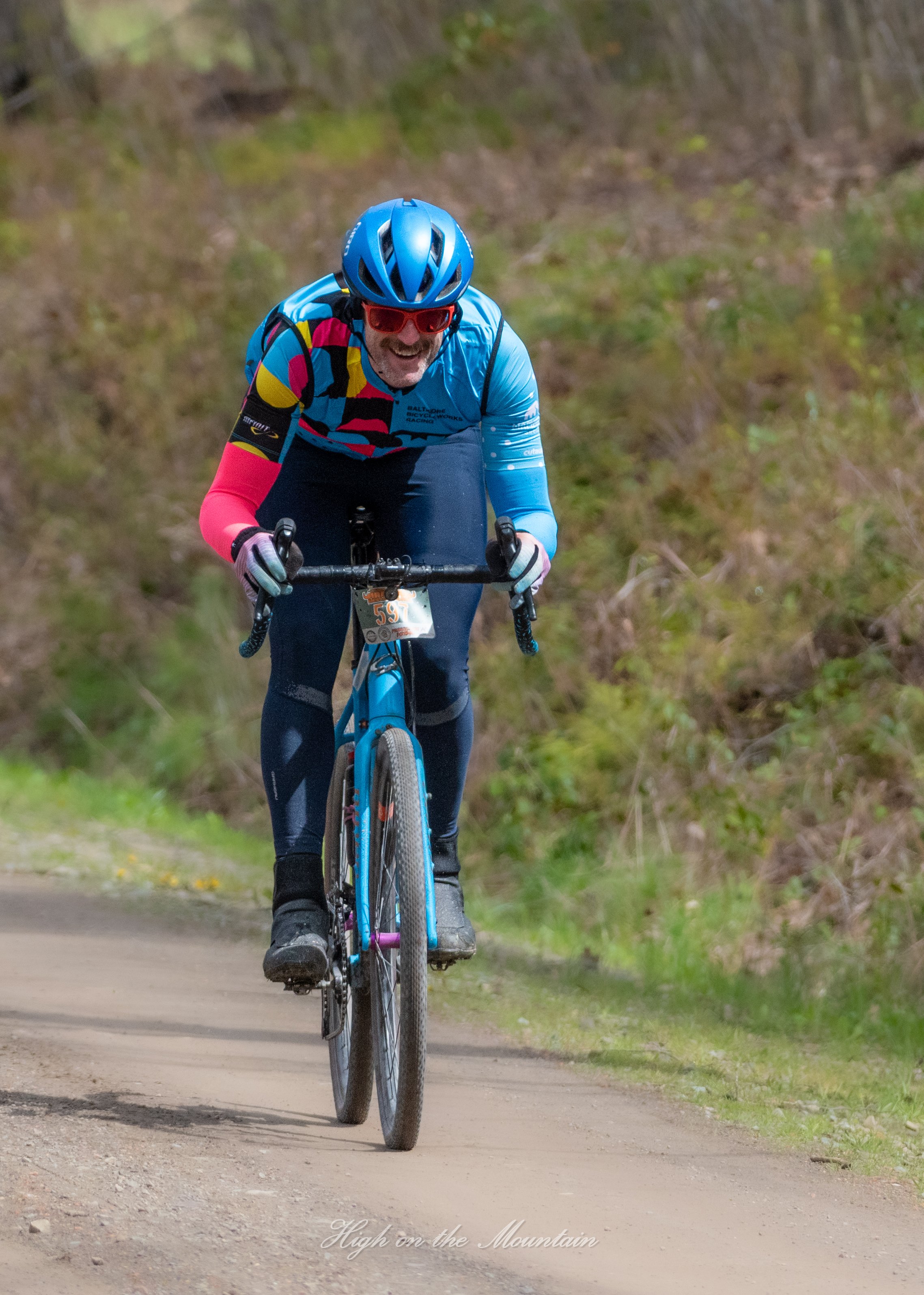 A man riding a mountain bike on a dirt trail in a wooded area, wearing a blue helmet, red sunglasses, a colorful cycling jersey, and black cycling pants.