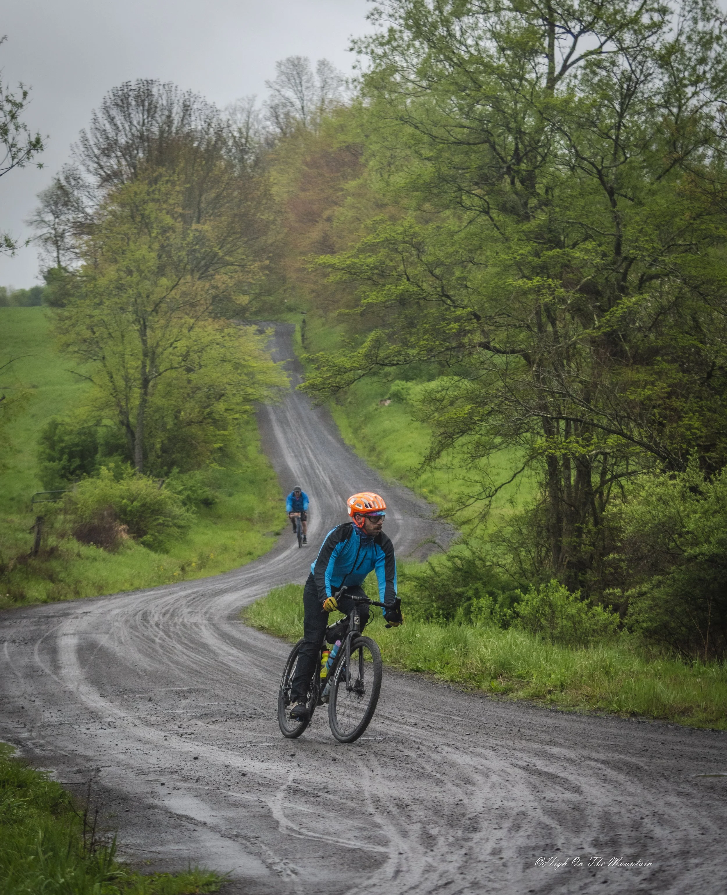 Cyclists riding on a winding dirt trail through a lush green forest, with trees and cloudy sky in the background.