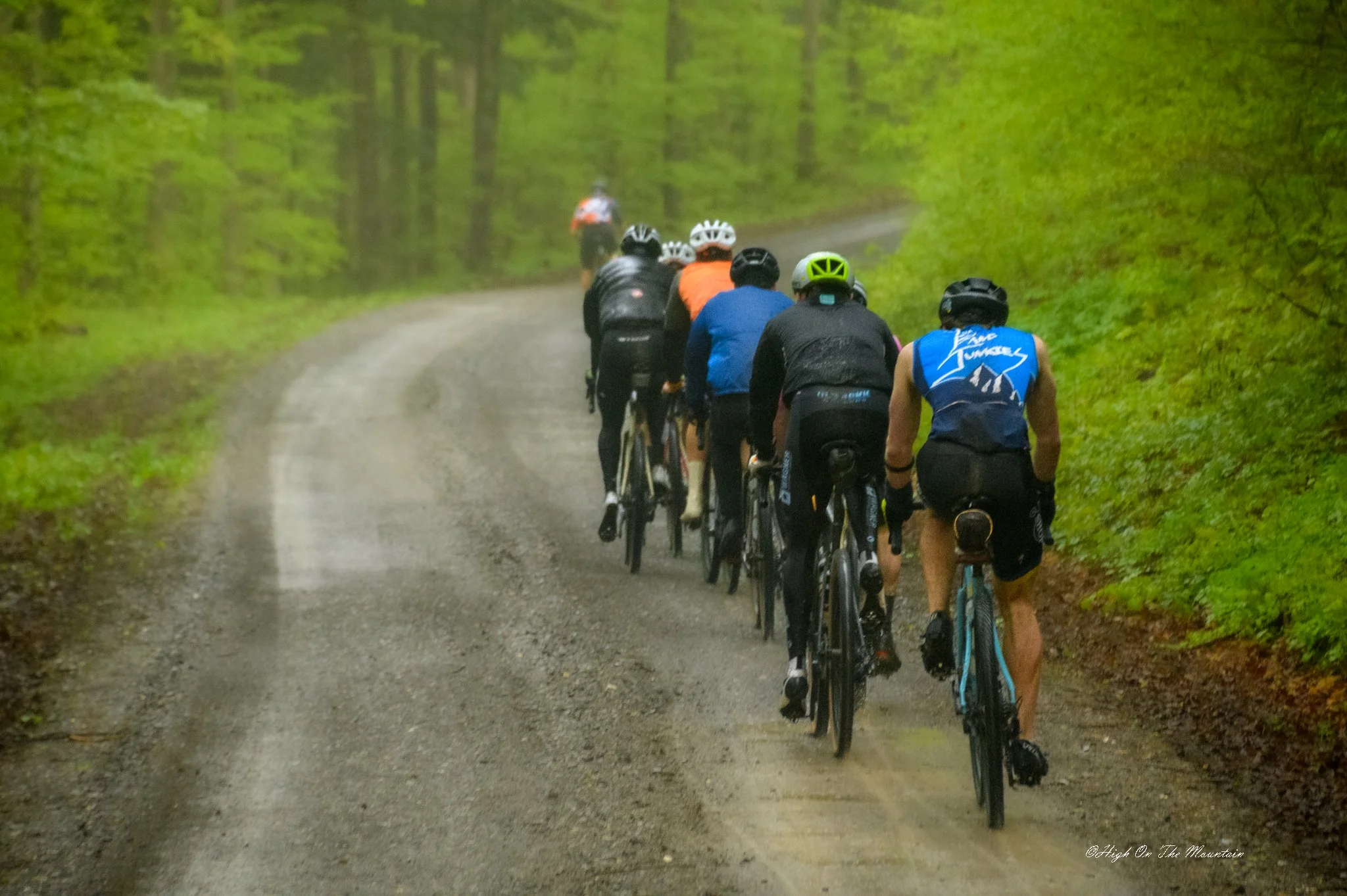 A group of cyclists wearing helmets and athletic gear riding mountain bikes on a dirt trail through a lush green forest.
