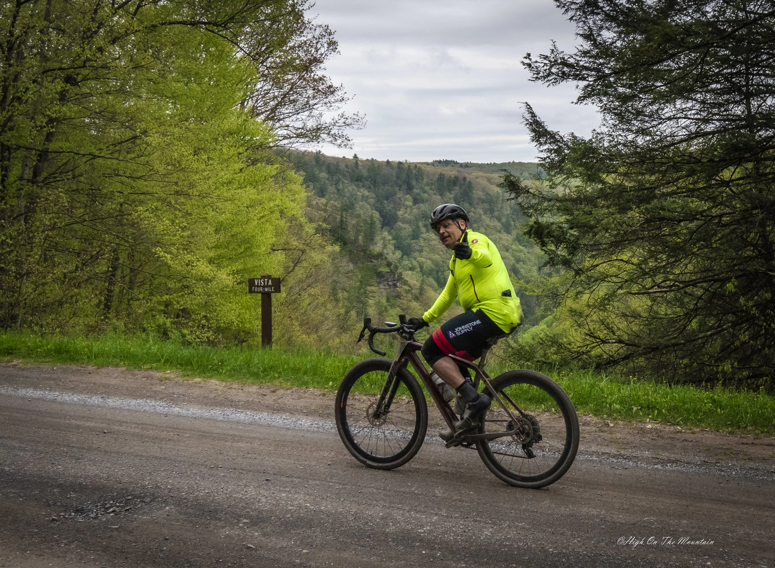 A cyclist in a bright yellow jacket riding on a gravel road surrounded by green trees, with a hillside in the background, giving a thumbs-up gesture.