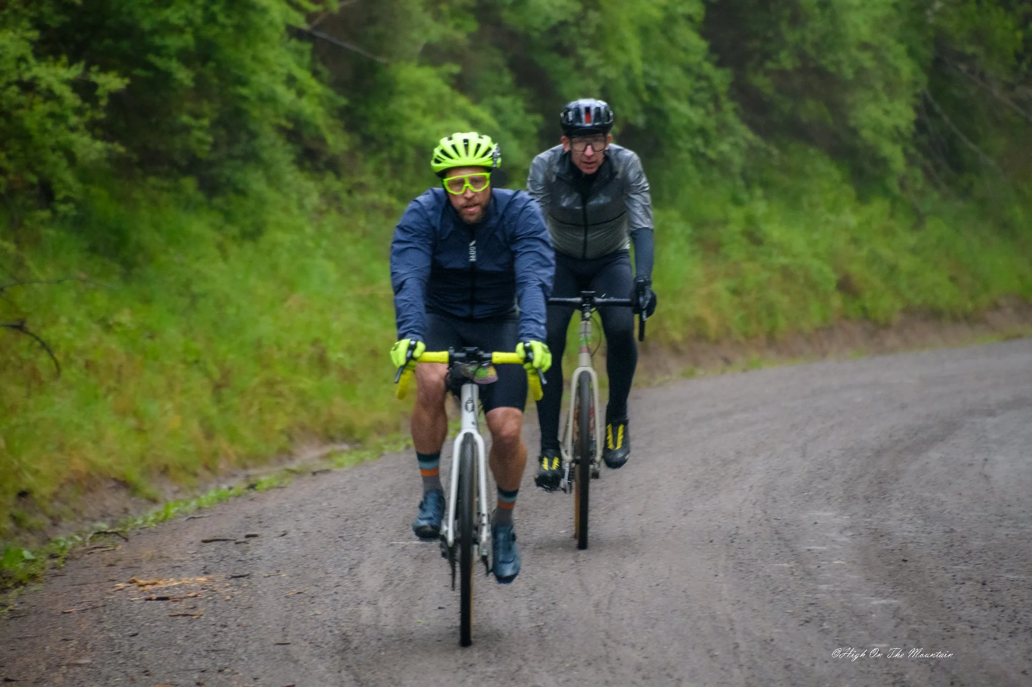 Two men riding bikes on a trail through a green forest, wearing helmets and athletic clothing.