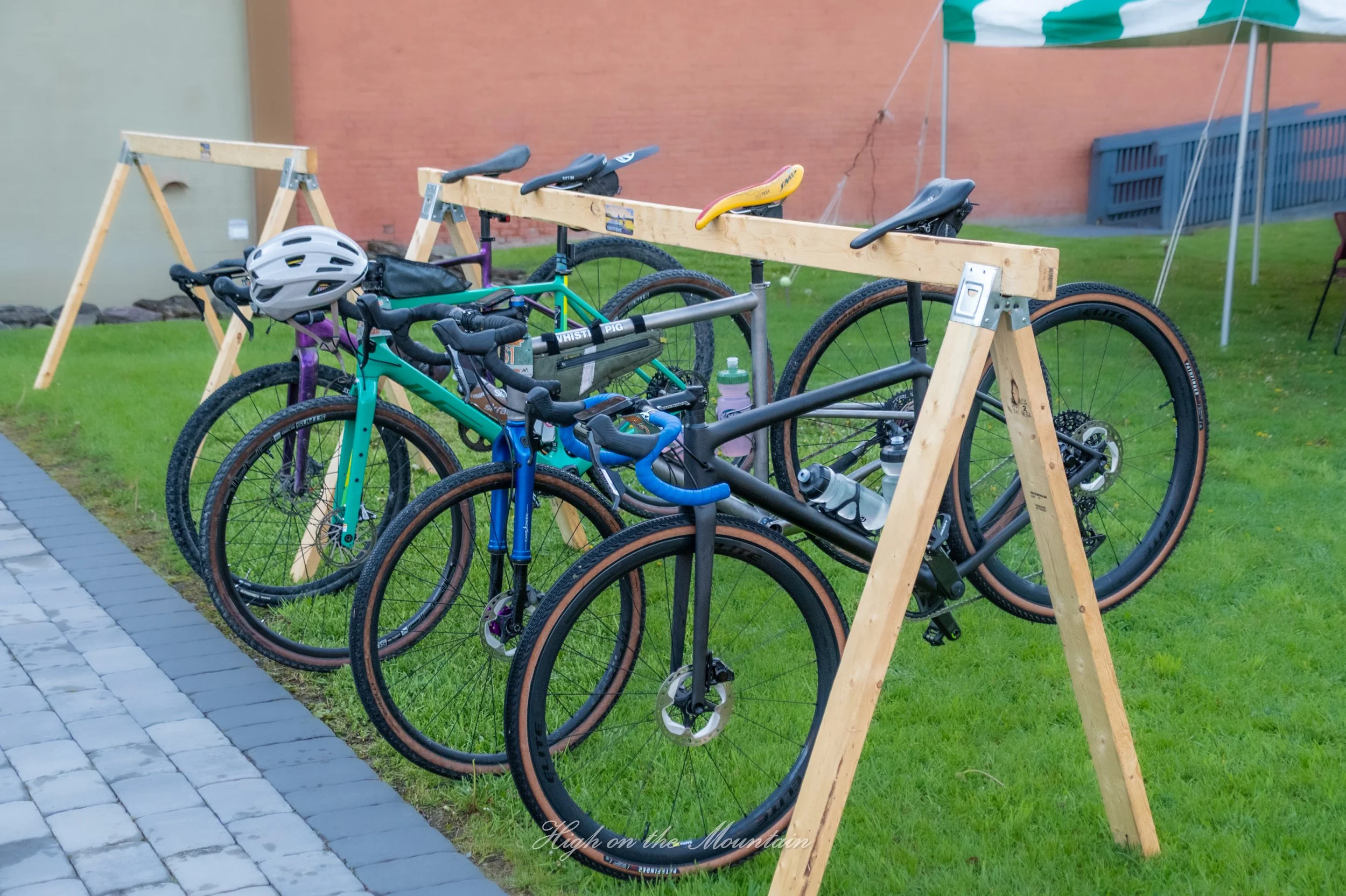 Multiple bicycles are mounted on a wooden bike rack outdoors, with a patch of green grass, a brick building, and a green and white striped umbrella in the background.