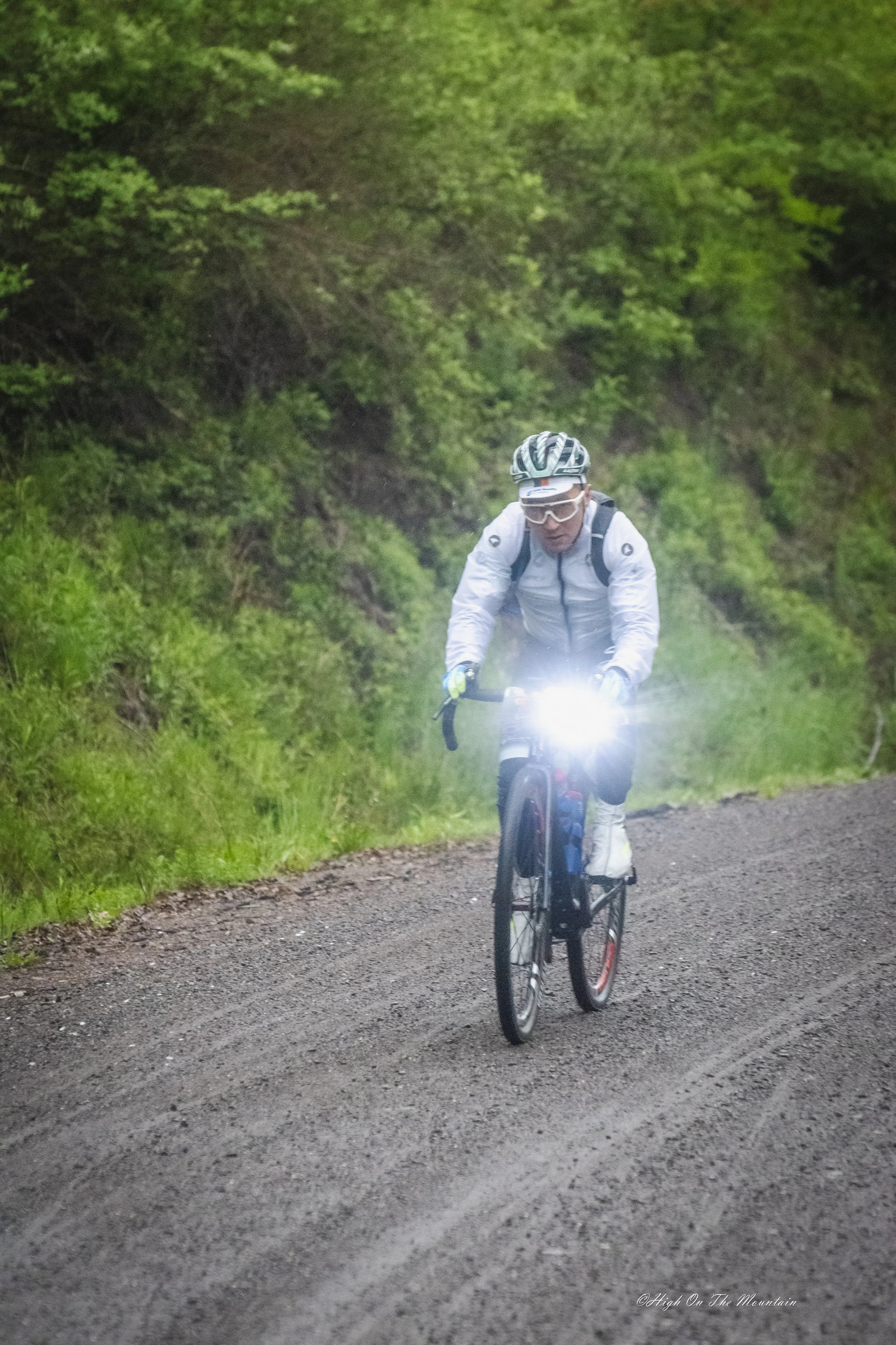 A man wearing a white jacket, helmet, and glasses rides a mountain bike on a dirt trail in a green forest with lush trees.