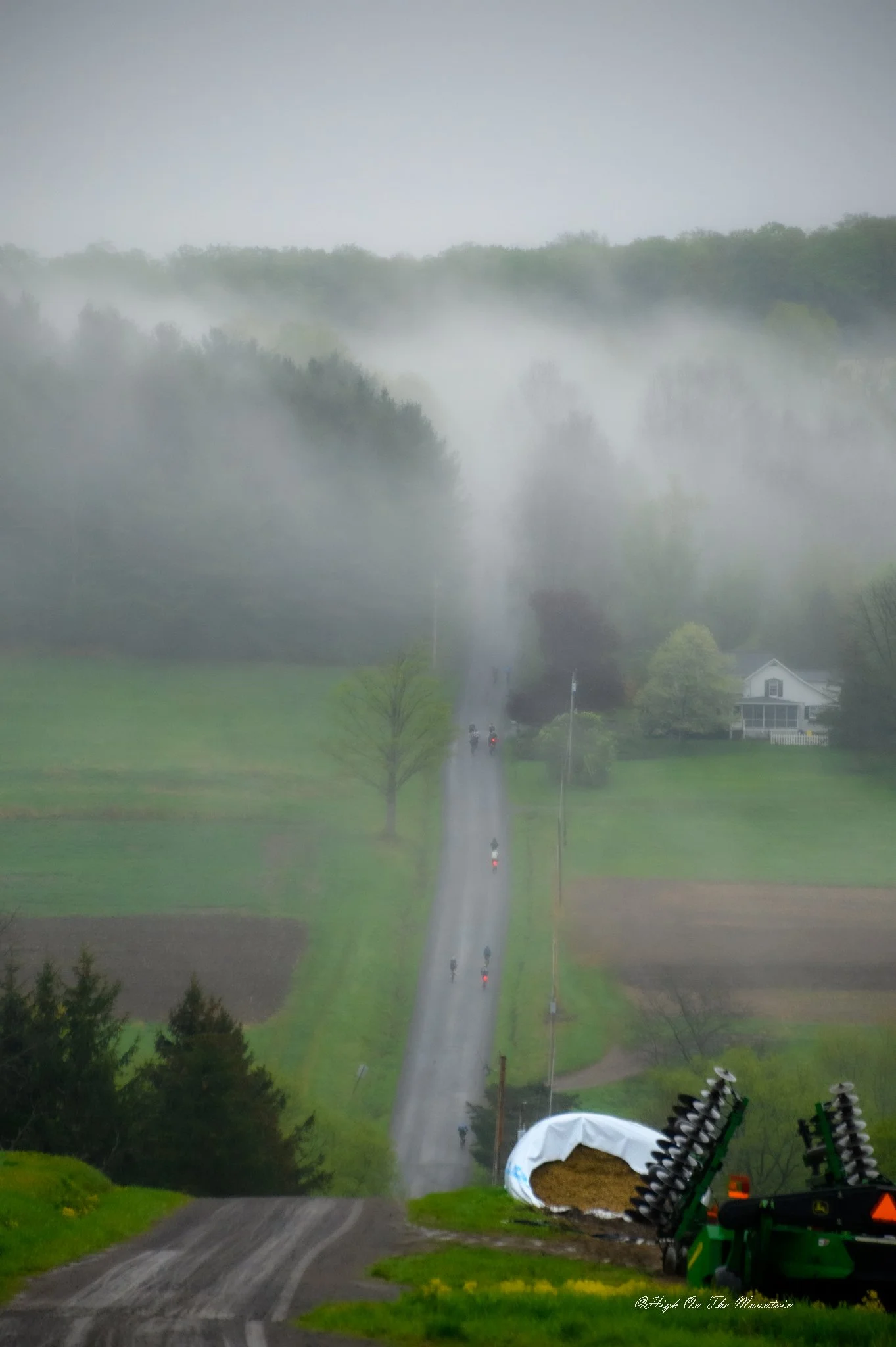 A rural landscape with a misty forested hillside in the background, a narrow paved road running through green fields, a few houses, and farm equipment in the foreground.
