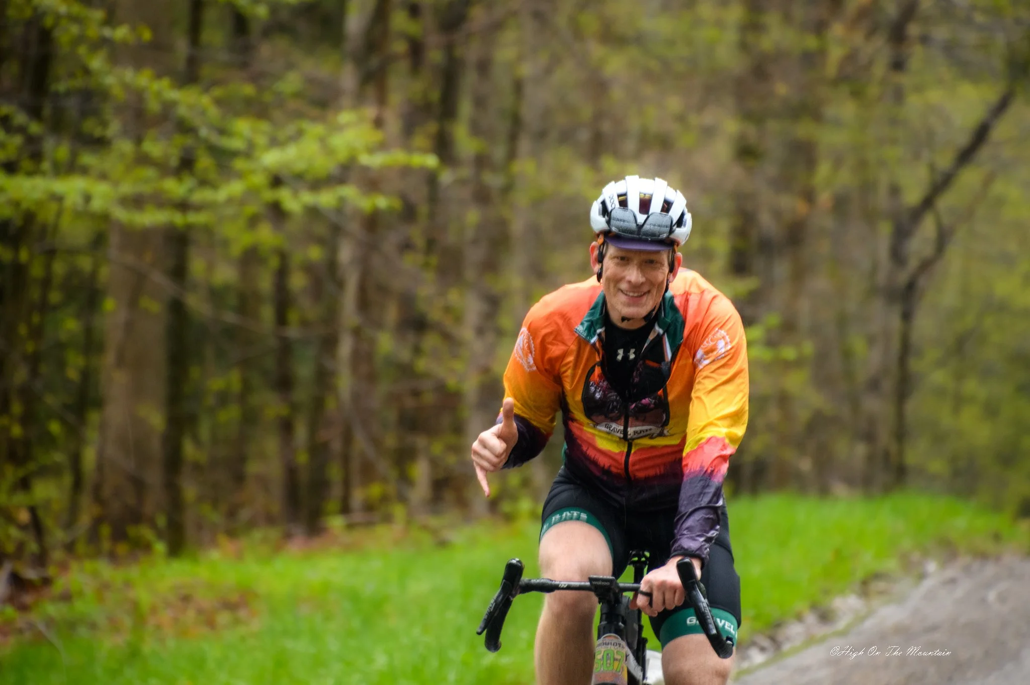 A man riding a mountain bike in a wooded area, wearing a helmet and an orange, rainbow, and green cycling jersey, smiling and giving a thumbs-up.