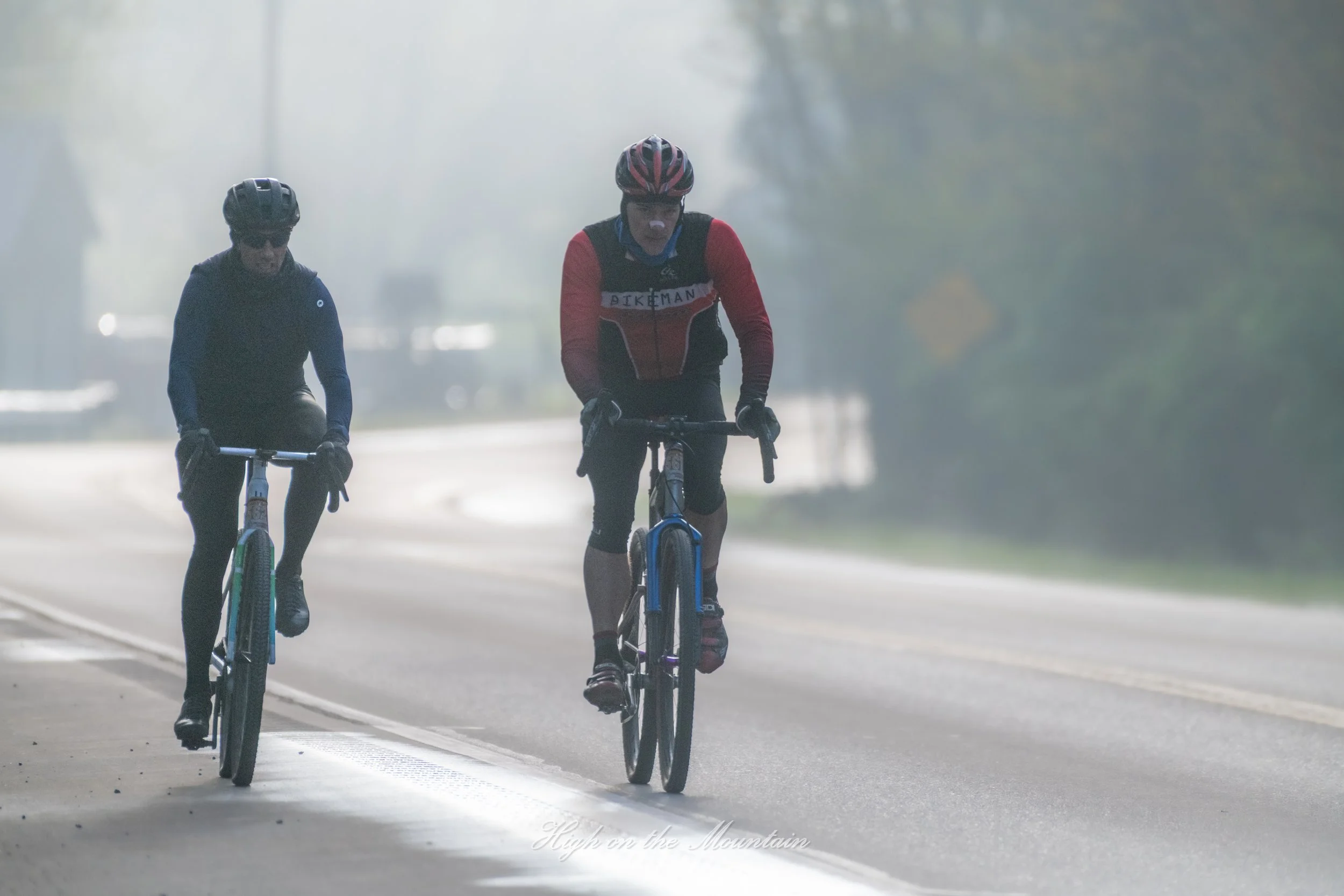 Two cyclists riding on a road, wearing helmets and athletic clothing, with a foggy background and trees.