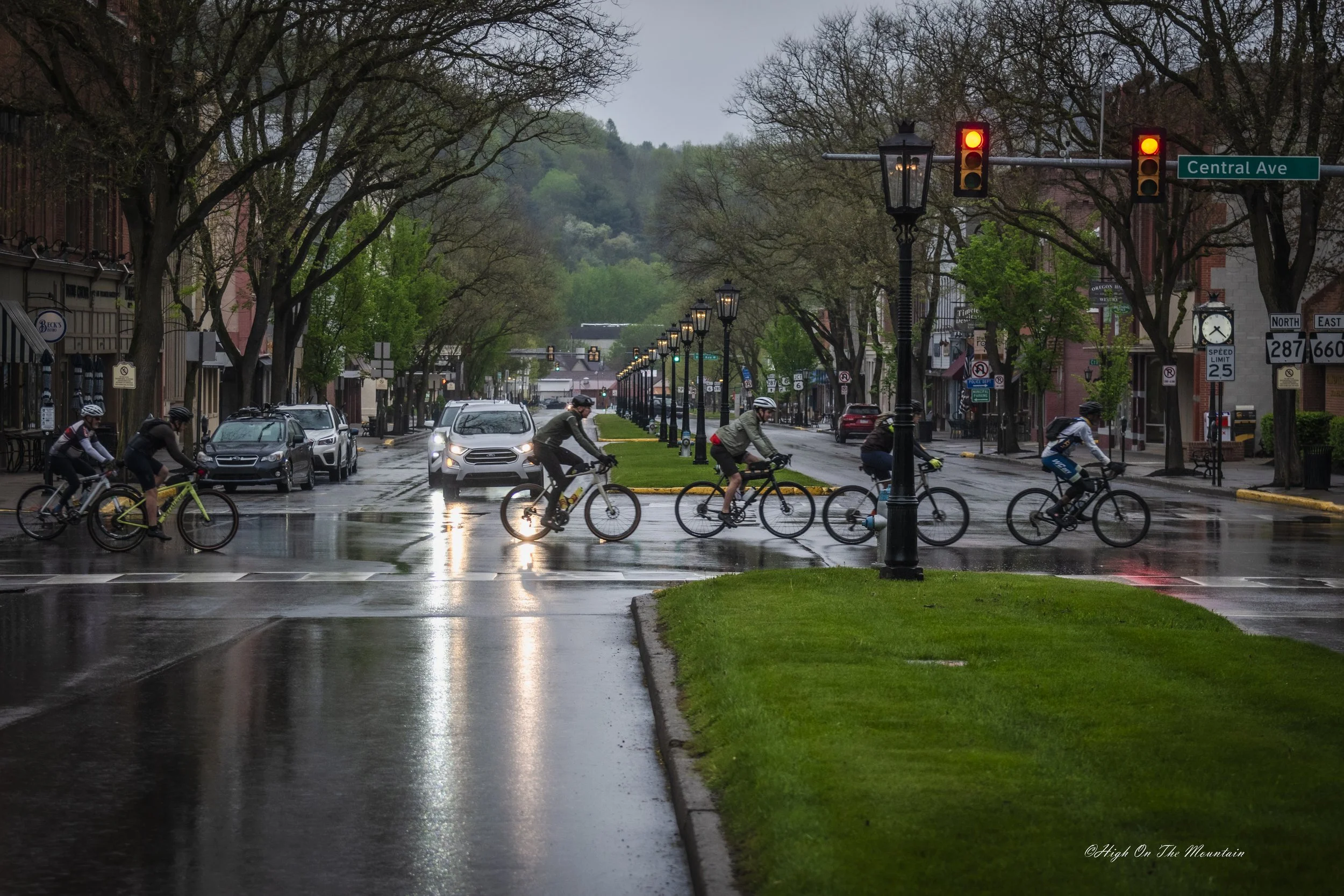 A rainy street scene with cyclists riding through a crosswalk, traffic lights showing red, and wet pavement with reflections. There are trees, lampposts, parked cars, and buildings lining the street.