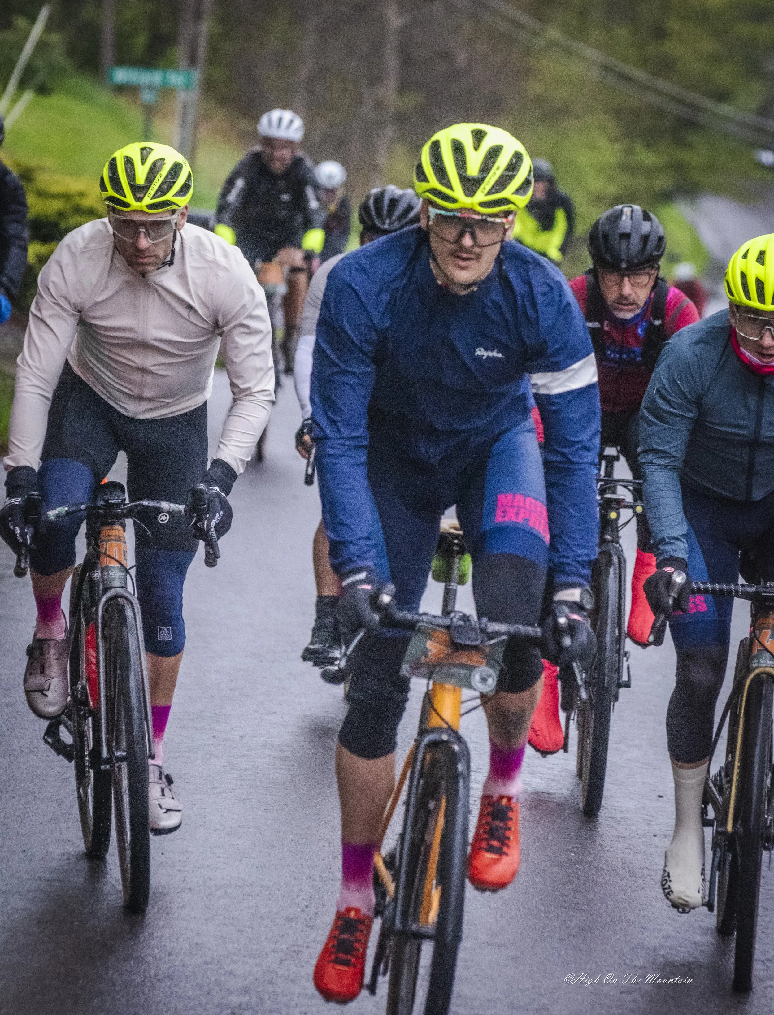 Group of cyclists riding on a wet road in a wooded area, wearing colorful biking gear and helmets.