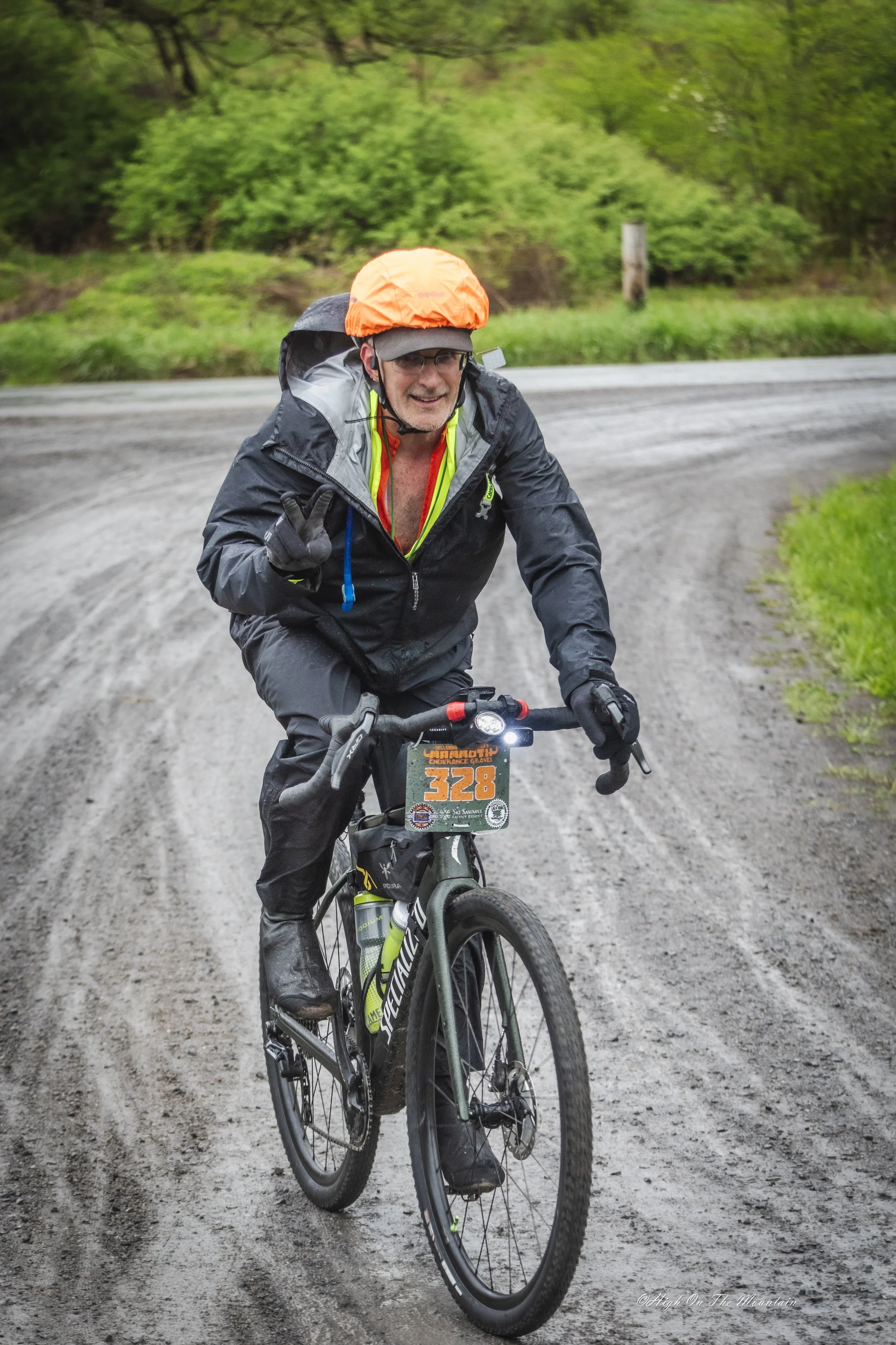 A man in a black jacket, glasses, and a helmet is riding a mountain bike on a dirt trail through a green wooded area. The man is smiling and making a peace sign with his right hand. There is a small amount of text on the front of the bike, including 